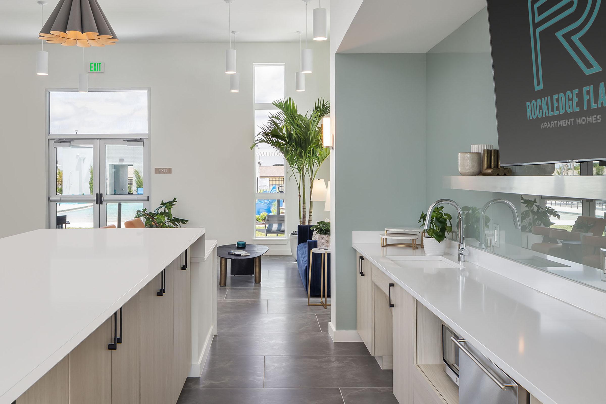 Interior view of a modern apartment lobby featuring a sleek white countertop, light wood cabinetry, and plants. Large windows allow natural light, with a glimpse of a pool area outside. Contemporary lighting hangs from the ceiling, adding a stylish touch to the space.