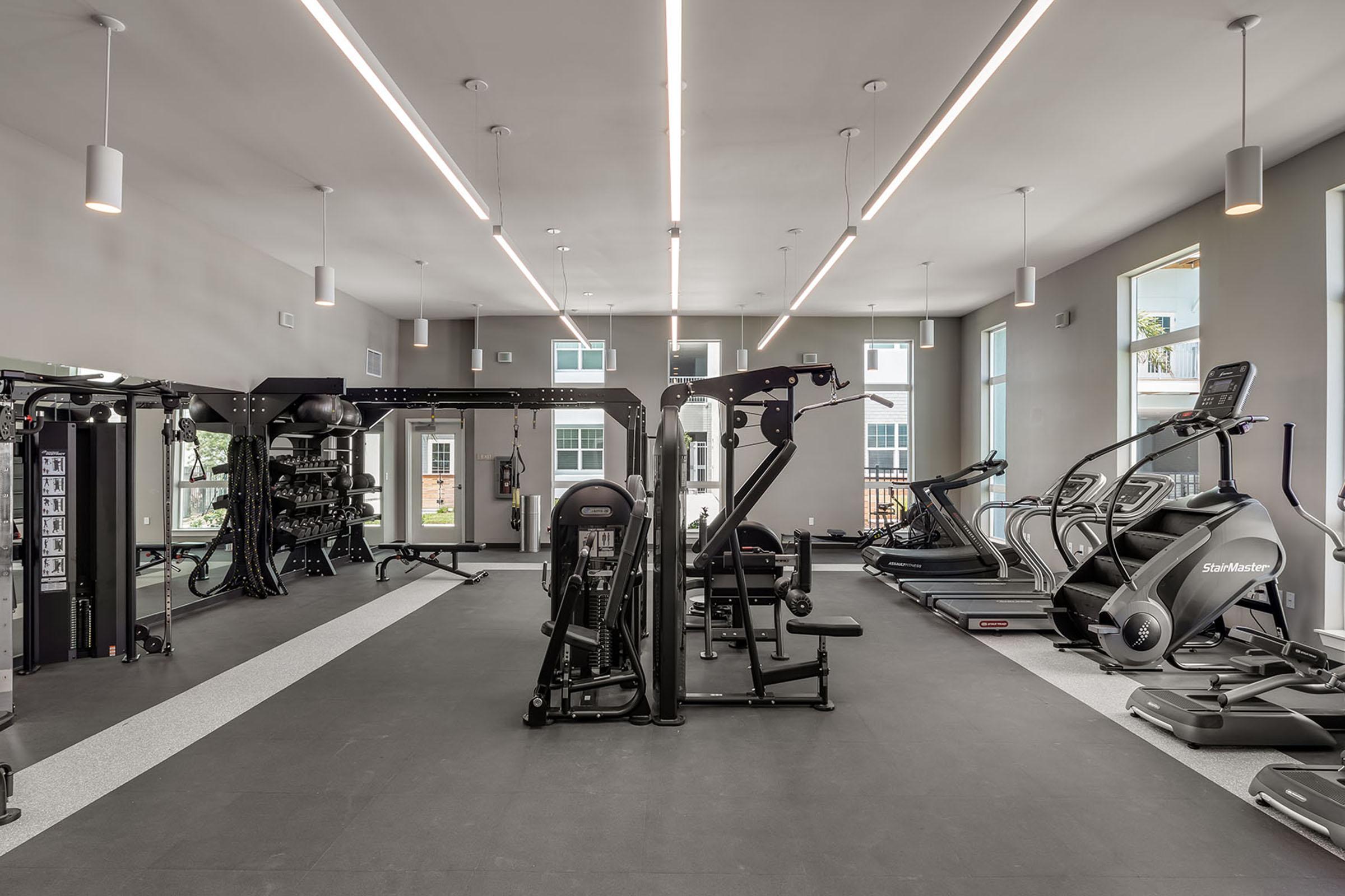 A modern gym interior featuring various exercise equipment, including weight machines, cardio machines, and an elliptical. The space is well-lit with overhead lights and large windows, creating an open and inviting atmosphere for workouts. The flooring is dark with a rubberized surface for safety.