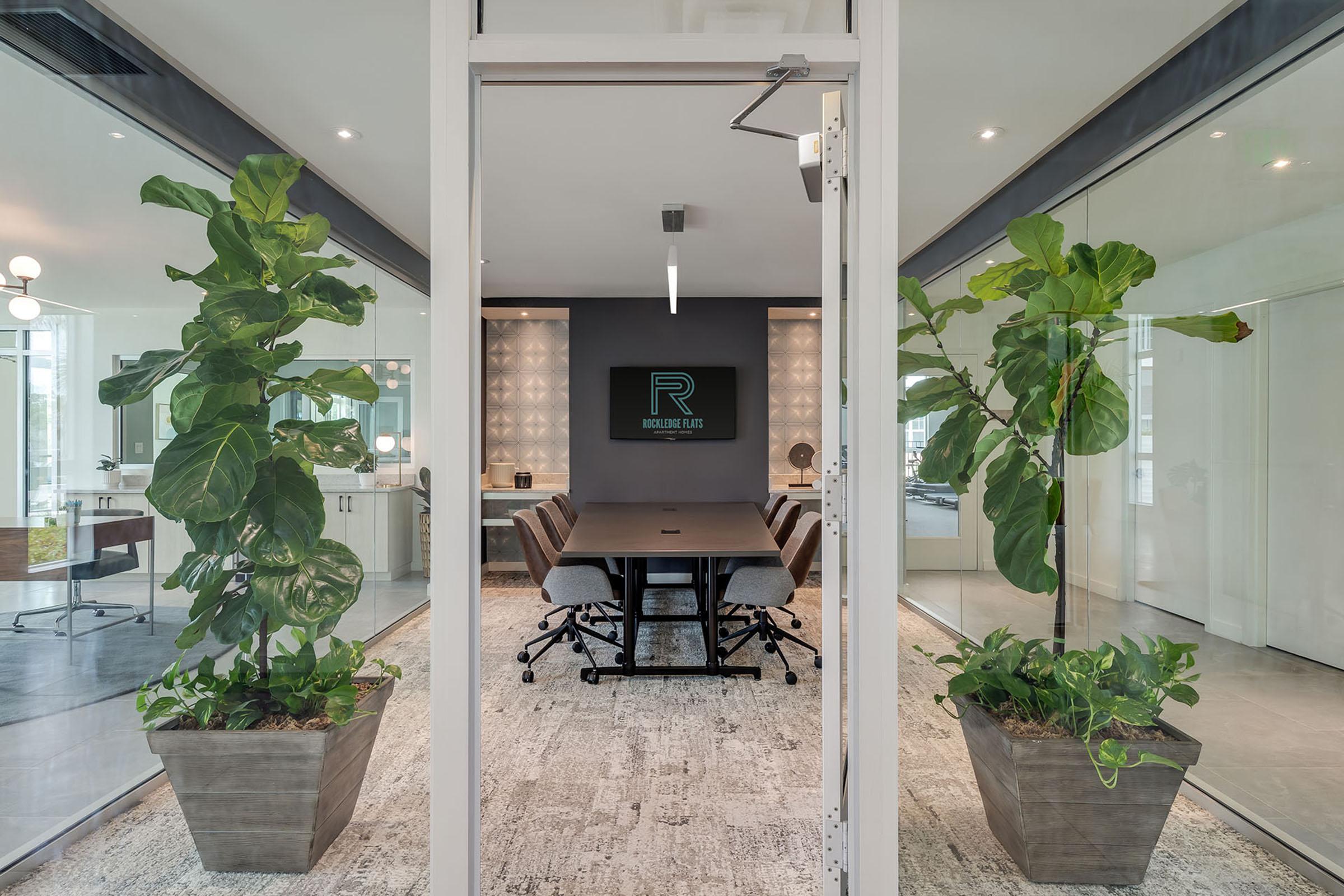 A modern conference room with glass walls featuring a wooden table surrounded by ergonomic chairs. Two large potted plants are positioned at the entrance, and a logo on the wall adds a professional touch. The room is well-lit with contemporary fixtures and a stylish carpet.
