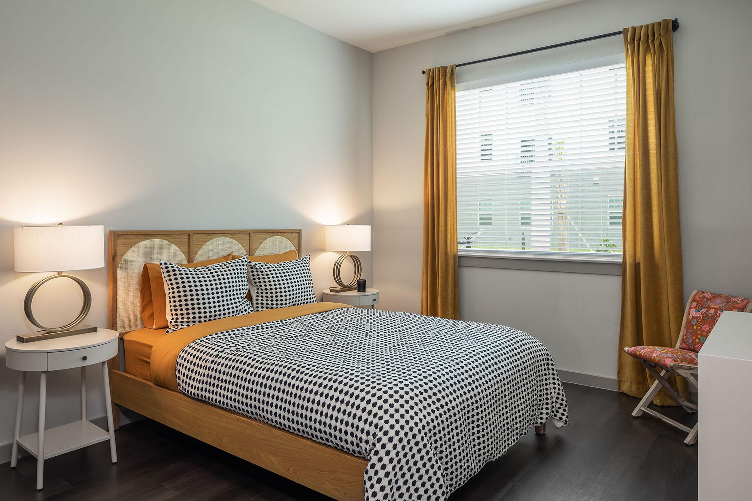A cozy bedroom featuring a bed with a polka dot patterned duvet and orange accent pillows. There are two modern bedside lamps on white tables, a window with sheer blinds letting in natural light, and an orange and white patterned chair in the corner, creating a warm and inviting atmosphere.