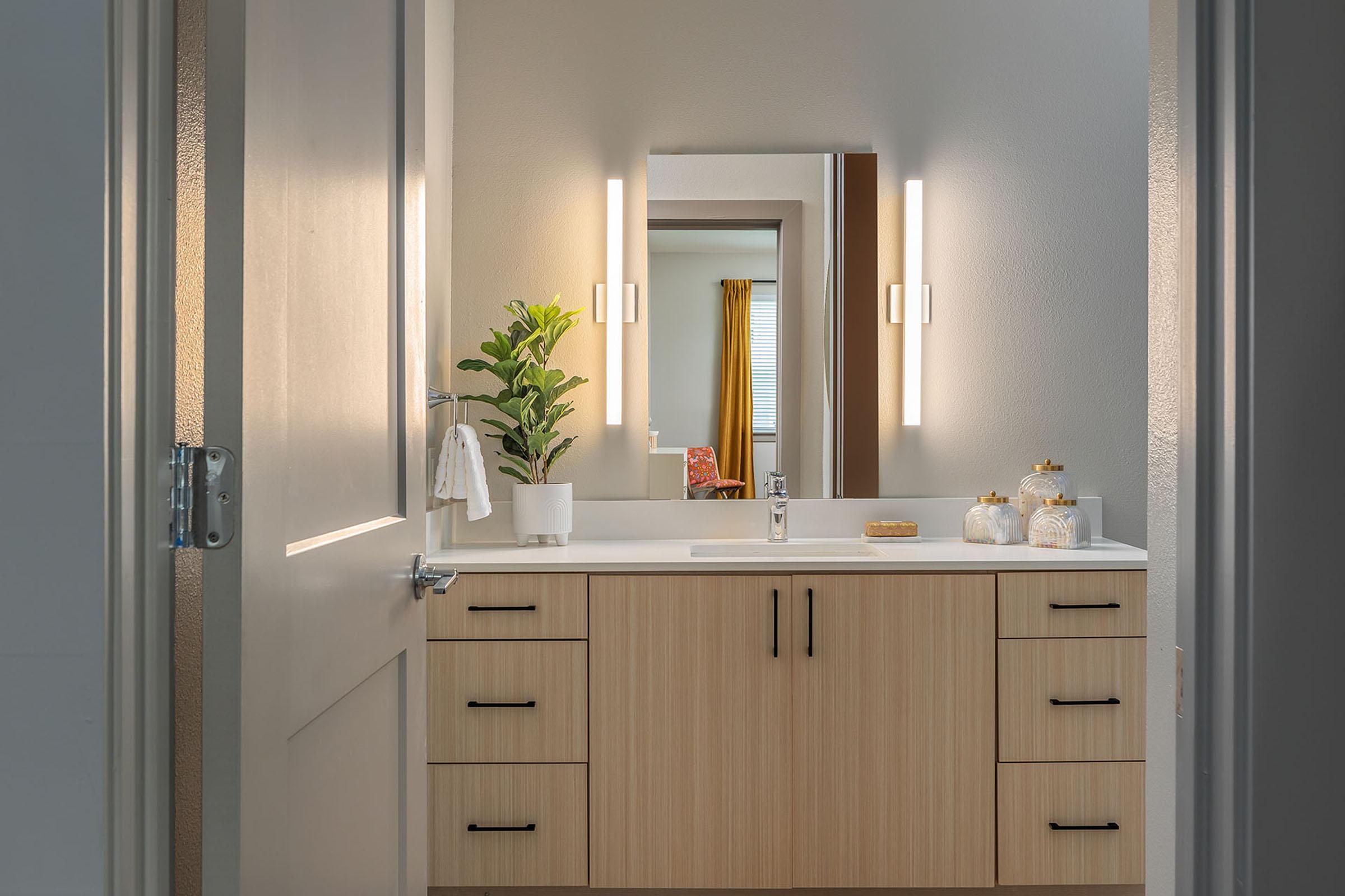 A modern bathroom with a light wooden vanity featuring drawers and a sink. Above the sink, there are two vertical light fixtures and a mirror. A small potted plant decorates the countertop, alongside decorative jars. The walls are painted in neutral tones, and a door partially opens to reveal the space.
