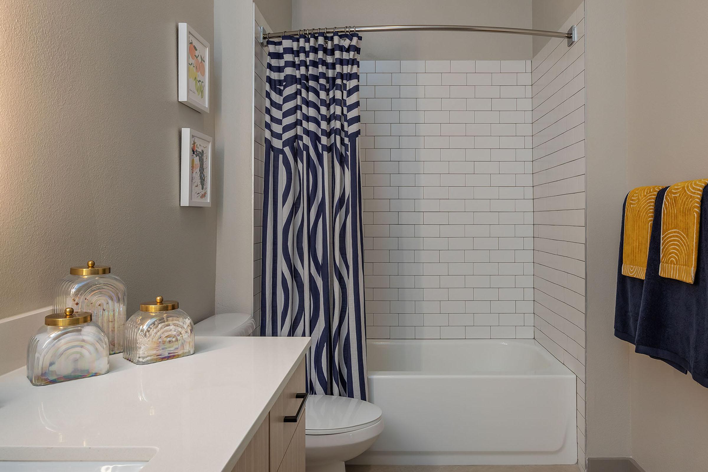 A modern bathroom featuring a white bathtub with a shower curtain in navy blue and white stripes. There are two framed pictures on the wall and a white countertop with decorative jars. A navy blue towel and a patterned yellow towel hang nearby, adding color to the clean, neutral space.