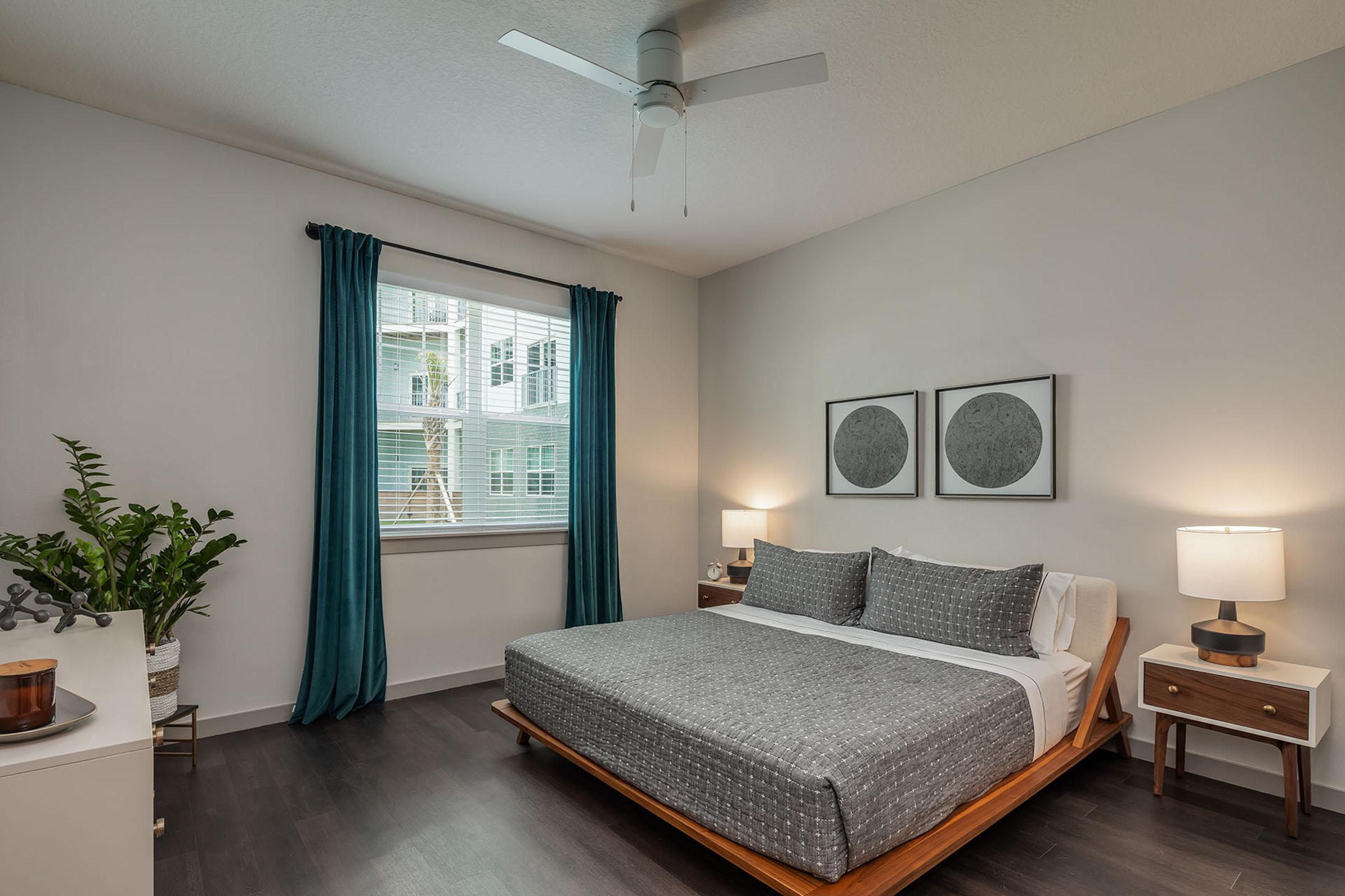 A modern bedroom featuring a large bed with gray bedding, a stylish wooden bed frame, and two bedside tables with lamps. The room has a large window adorned with teal curtains, allowing natural light to brighten the space. A green plant is placed near a dresser, enhancing the room's aesthetic.