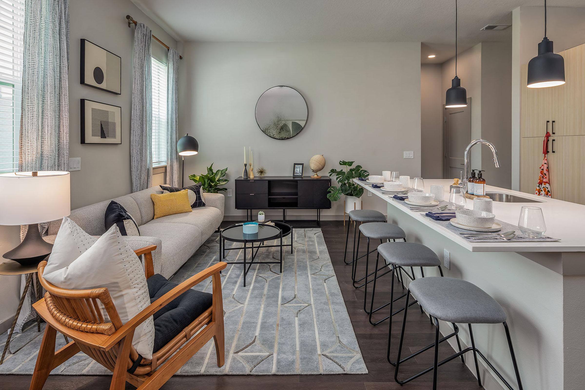 A modern, spacious living room with a light gray sofa and a round coffee table on a patterned rug. Nearby, a dining area with bar stools and a countertop is set with dishes. Large windows let in natural light, and decorative plants and artwork add warmth to the space.