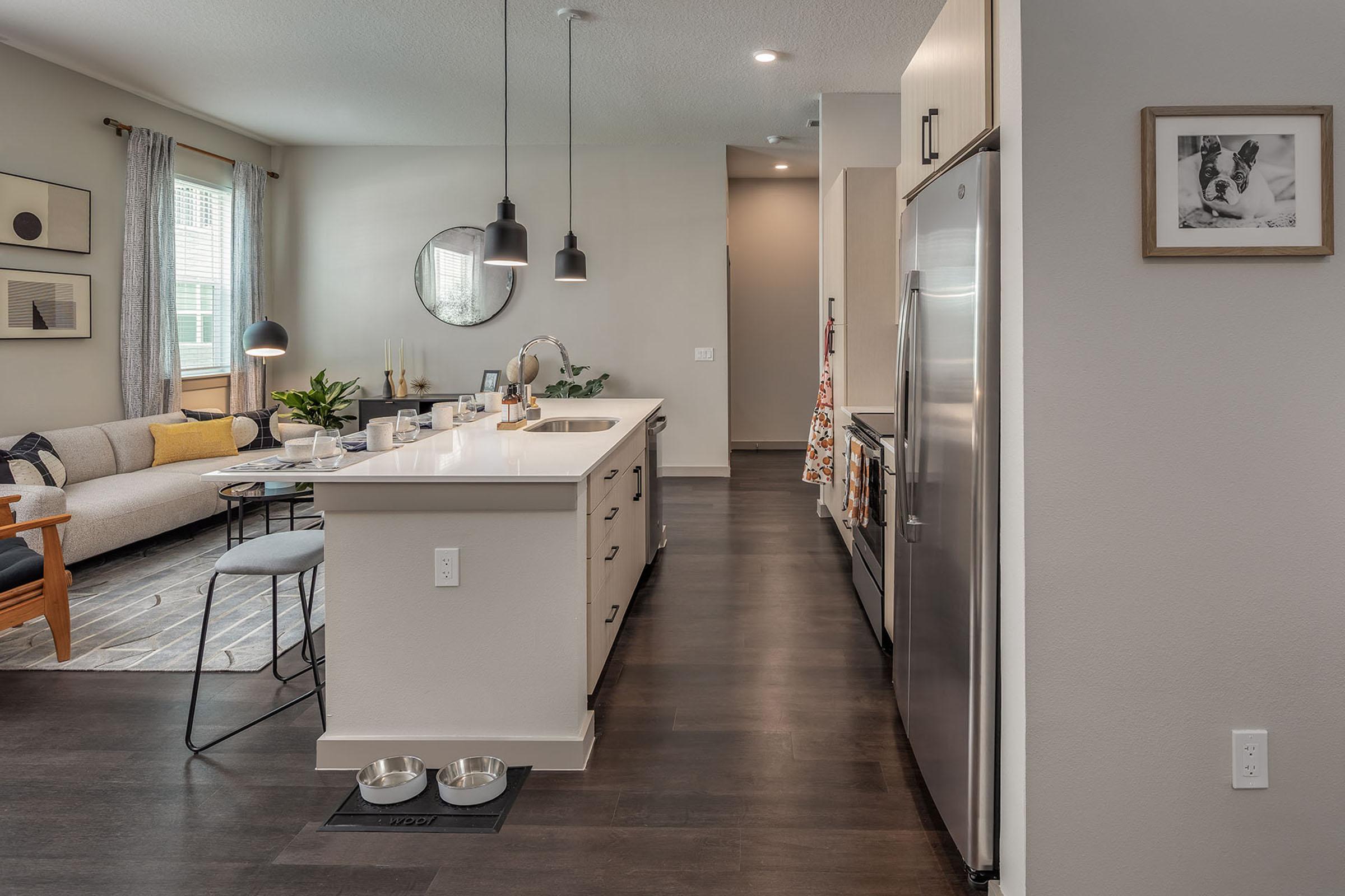Modern kitchen with an island, featuring stainless steel appliances and minimalistic decor. A cozy living area is visible in the background, with a sofa and a round mirror. Stylish lighting fixtures hang above the island, and there's a pet food bowl on the floor. The color scheme is neutral and inviting.