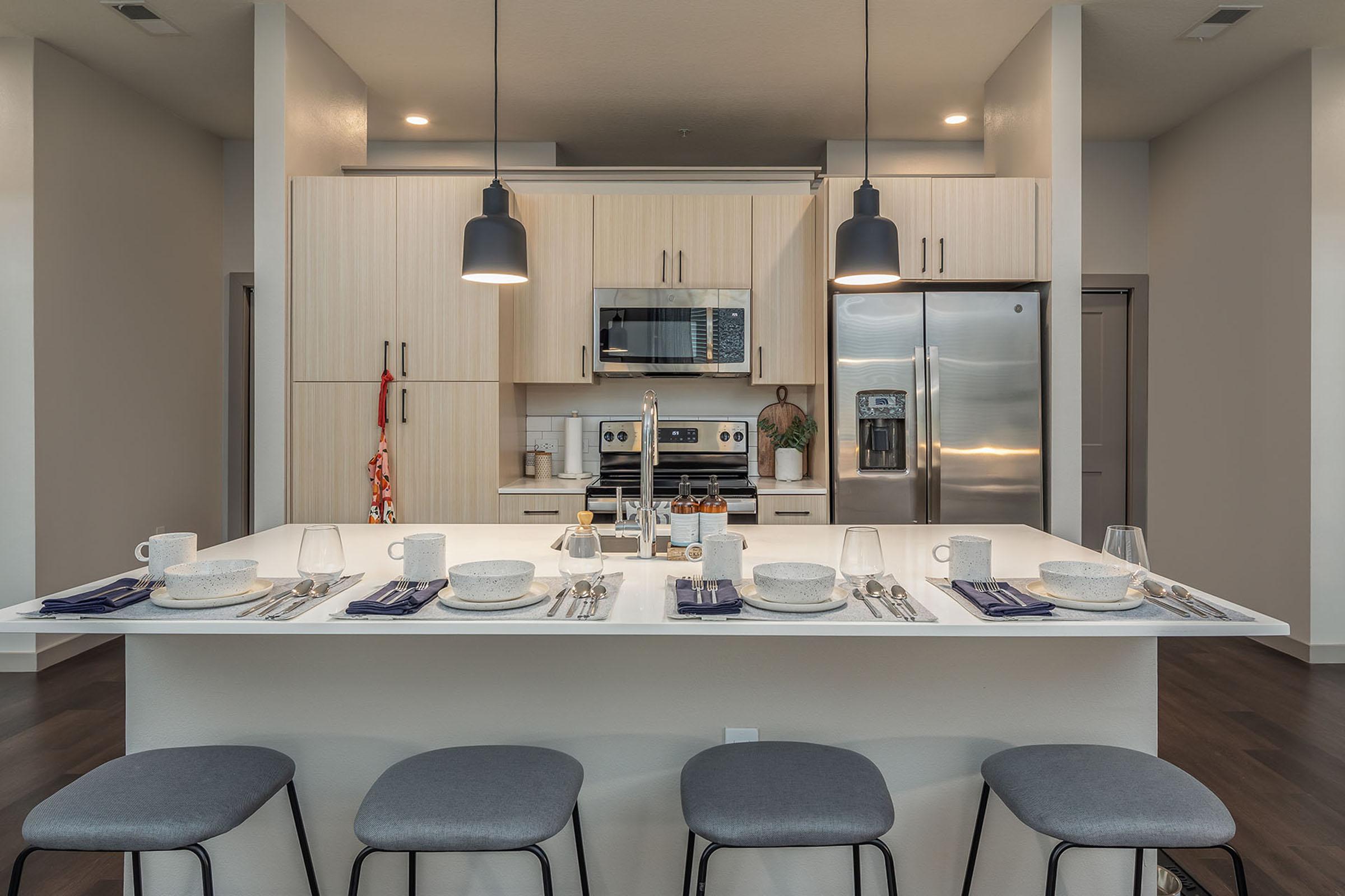 Modern kitchen with light wood cabinetry and stainless steel appliances. An island with a white countertop features a neatly set dining arrangement, including bowls, glasses, and utensils. Four gray bar stools are positioned around the island, while hanging pendant lights illuminate the space.