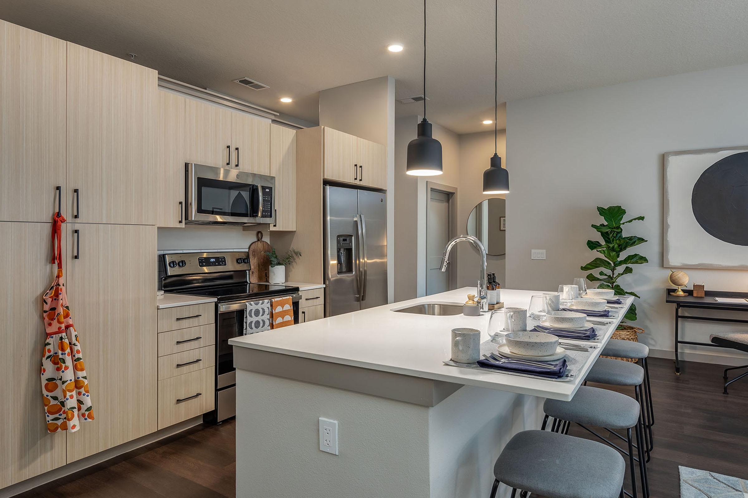 Modern kitchen featuring light wood cabinetry, stainless steel appliances, and a large island with bar stools. A dining set is arranged on the island with plates and cups, while a stylish plant adds a touch of greenery. Pendant lights illuminate the space, creating a warm ambiance.