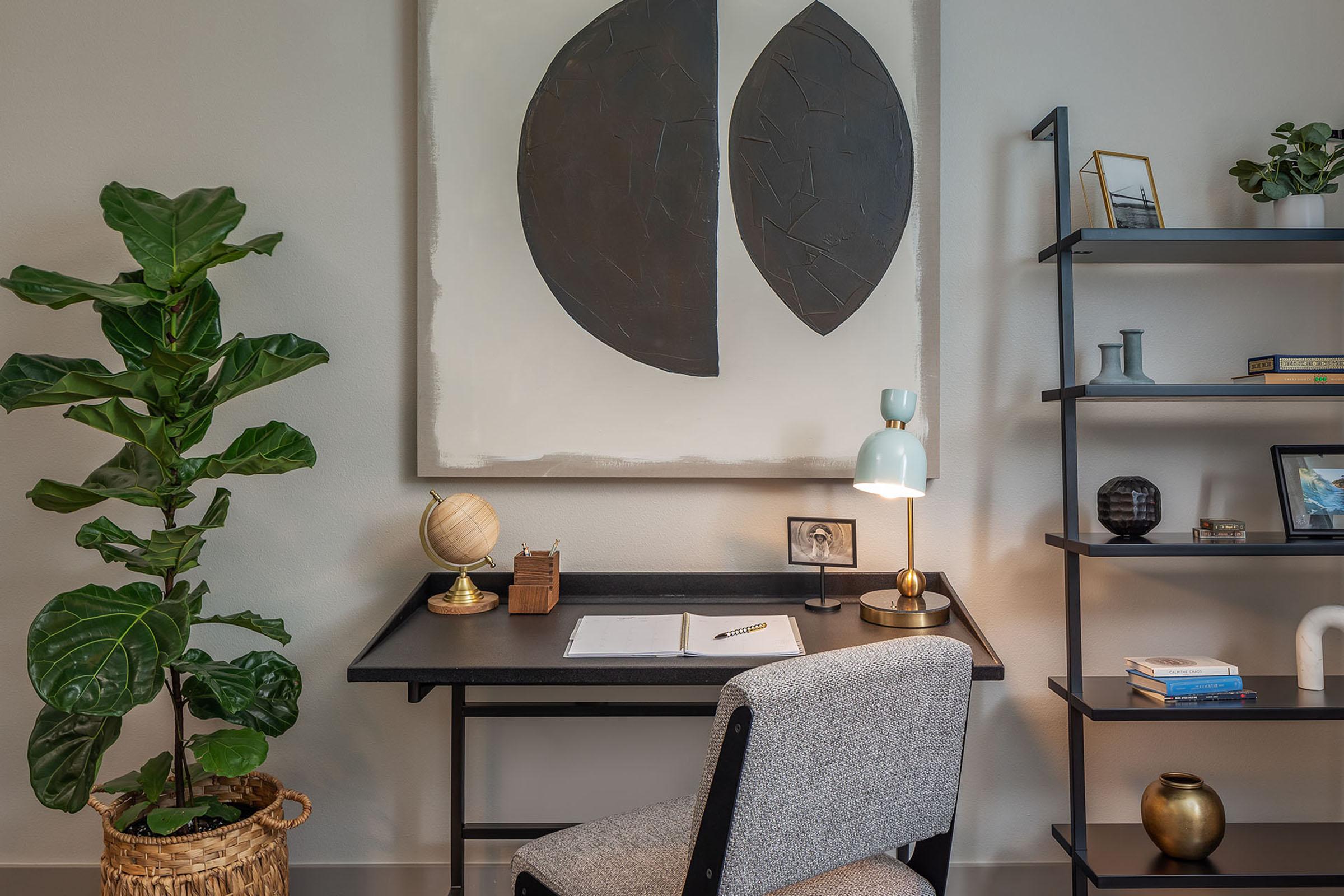 A stylish workspace featuring a black desk with a notebook and pen, a light blue lamp, and a globe. A large abstract painting with two black shapes hangs above. A tall green plant adds a touch of nature, while a black bookshelf holds various decorative items and books. Warm, modern decor creates a cozy atmosphere.