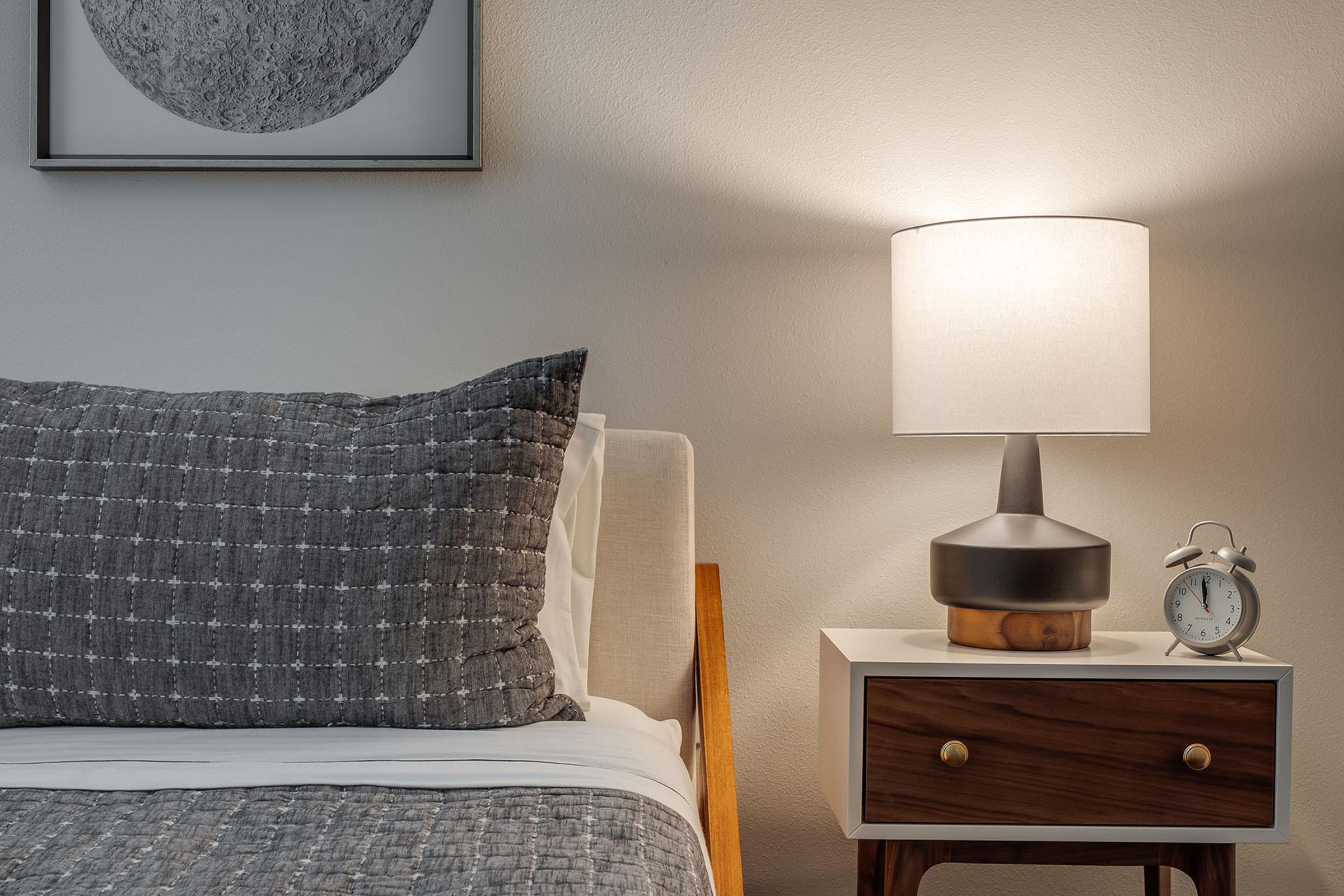 A cozy bedroom scene featuring a neatly made bed with a gray patterned throw pillow. Next to the bed, there's a modern bedside table with a round wooden lamp and an alarm clock, accompanied by a simple wall art piece depicting a circular design. The lighting creates a warm atmosphere.