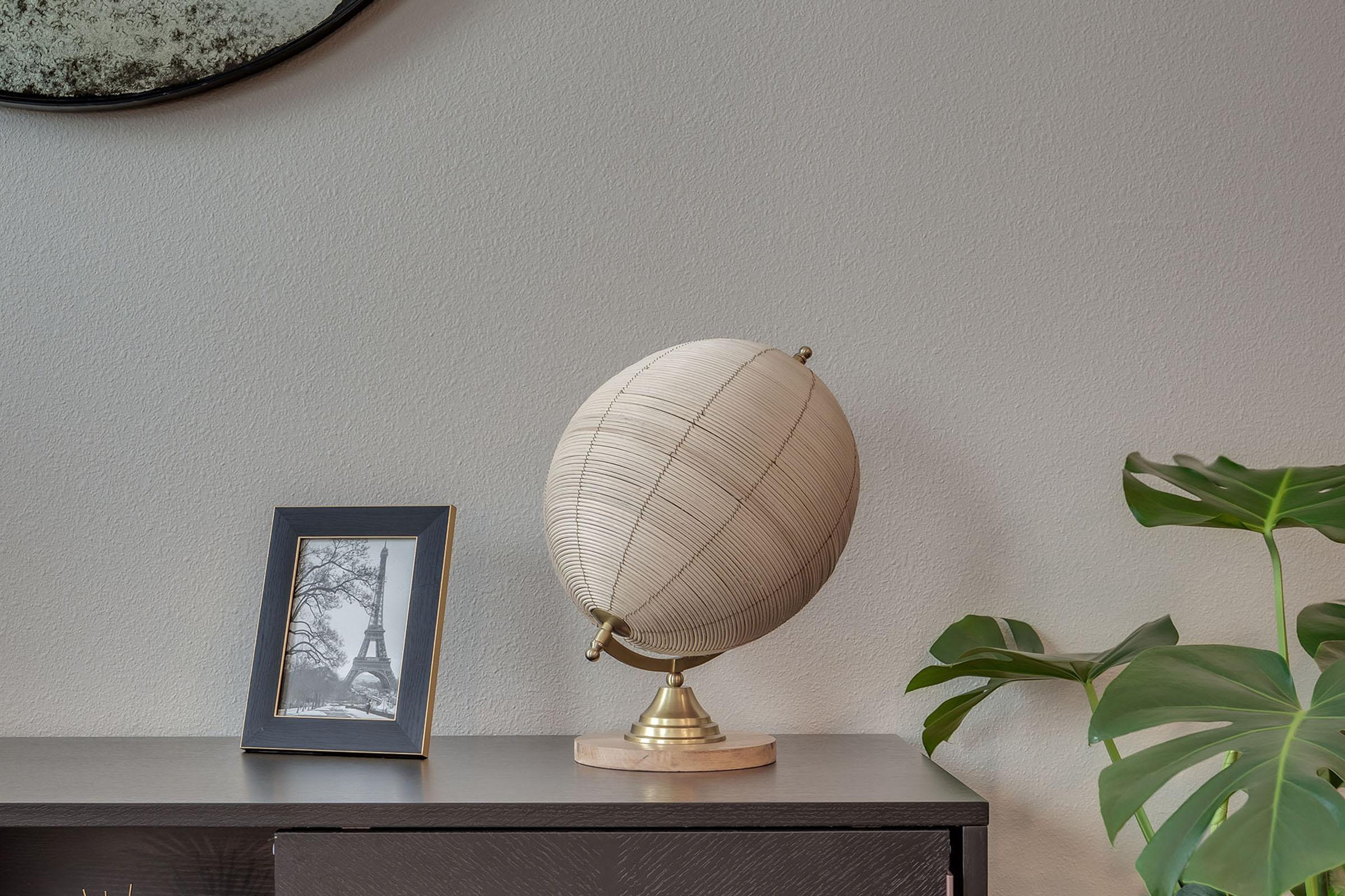 A decorative globe on a stand sits on a dark wooden surface, accompanied by a framed black-and-white photo of the Eiffel Tower and a lush green plant. A round mirror can be seen in the background against a light-colored wall.