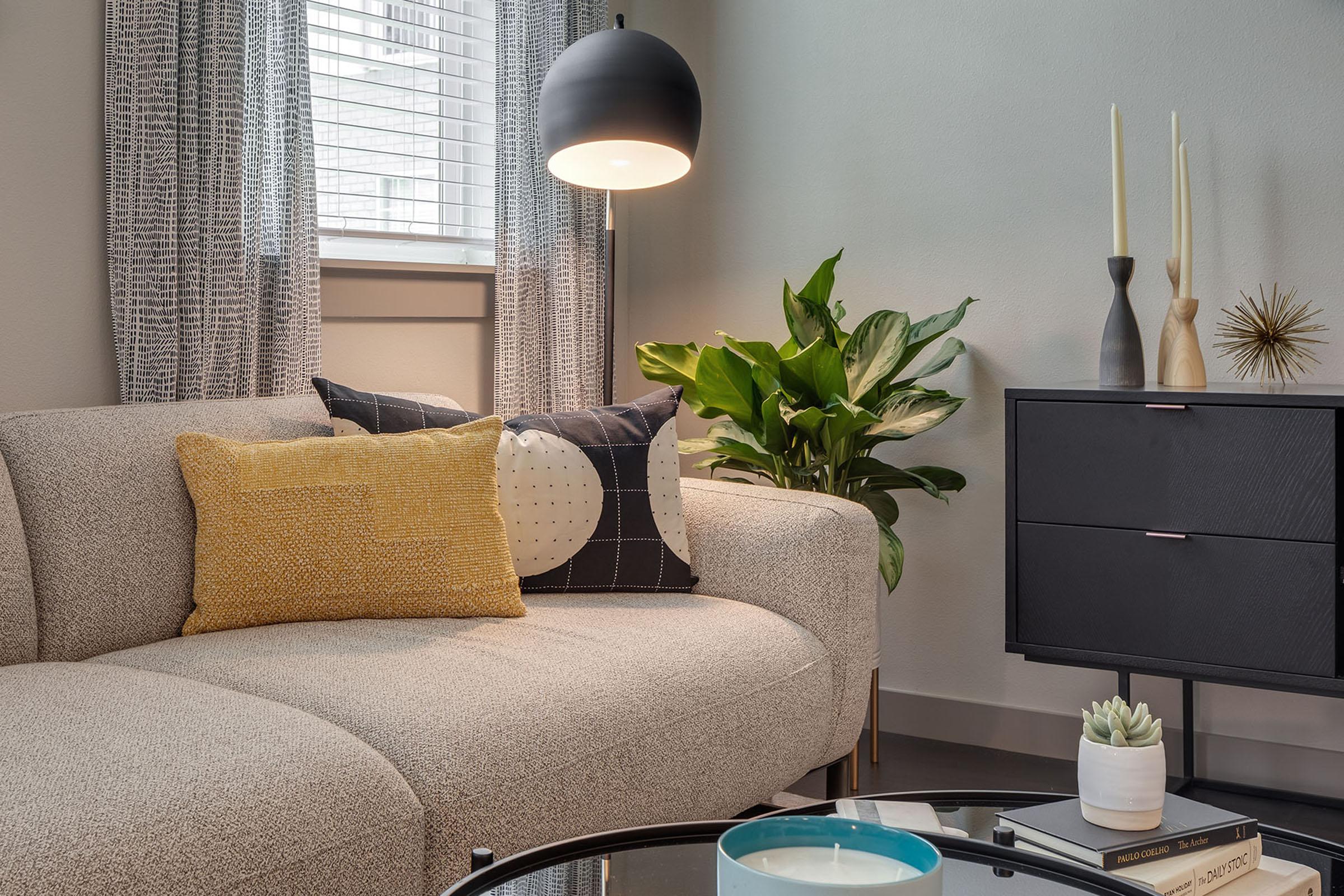 Modern living room featuring a light-colored curved sofa with patterned throw pillows, a sleek black side table with decorative items, a potted plant, and a floor lamp. Natural light filters through window blinds, creating a cozy atmosphere.