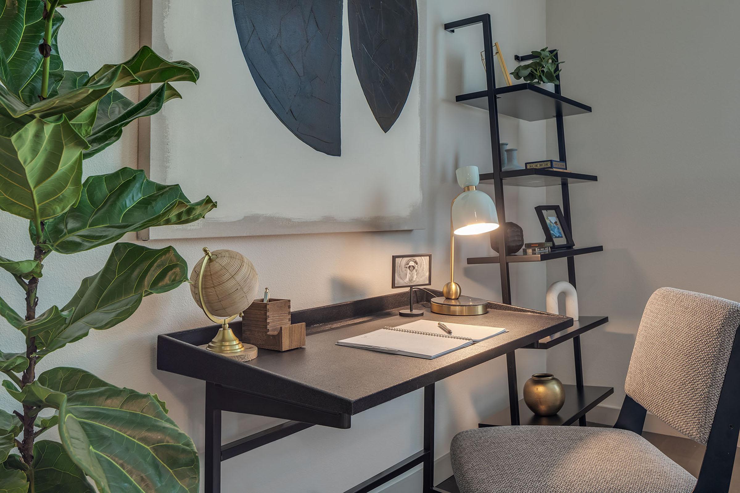 A modern workspace featuring a minimalist black desk with a decorative globe and notepad. A stylish lamp illuminates the area. Behind the desk is a leafy plant and a black shelving unit holding items like a picture frame and a decorative object. The overall aesthetic is sleek and inviting.