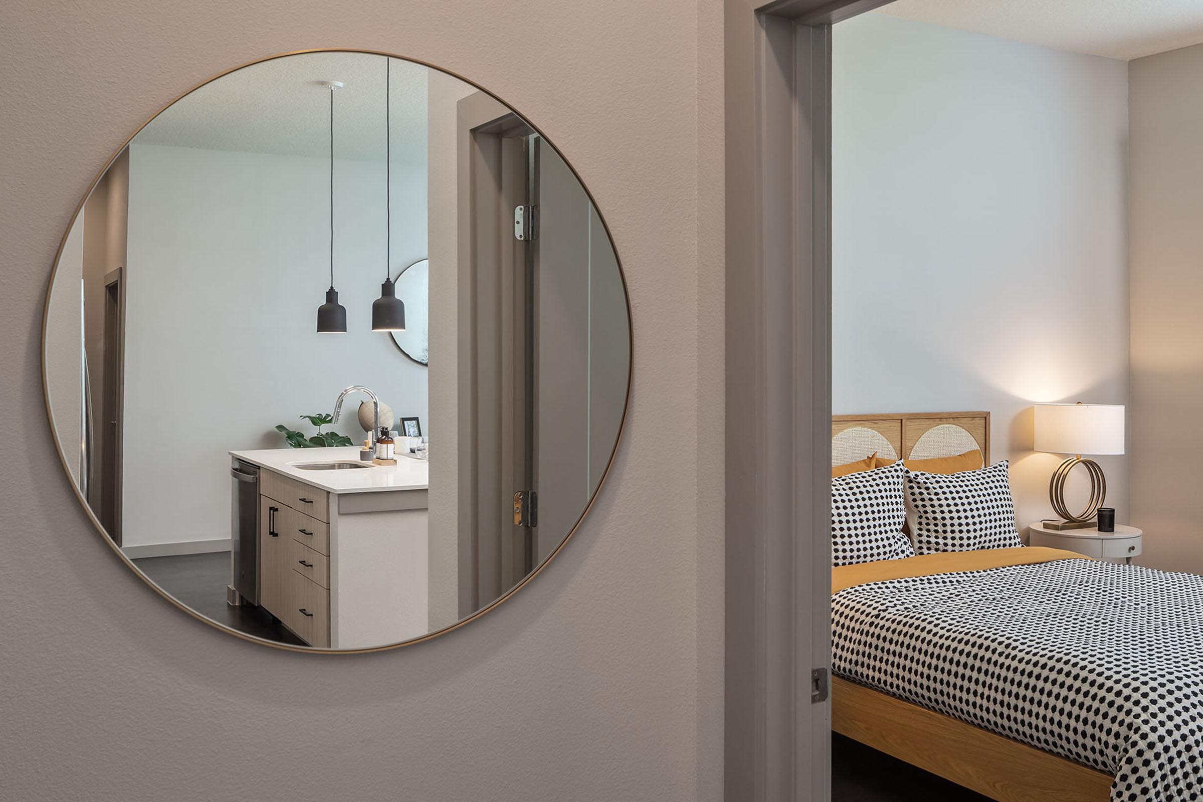 A modern bedroom visible through a round mirror, featuring a cozy bed with patterned bedding and a lamp on a nightstand. In the foreground, a stylish kitchen area is reflected in the mirror, showing minimalist decor and pendant lighting. The overall color scheme is neutral and contemporary.