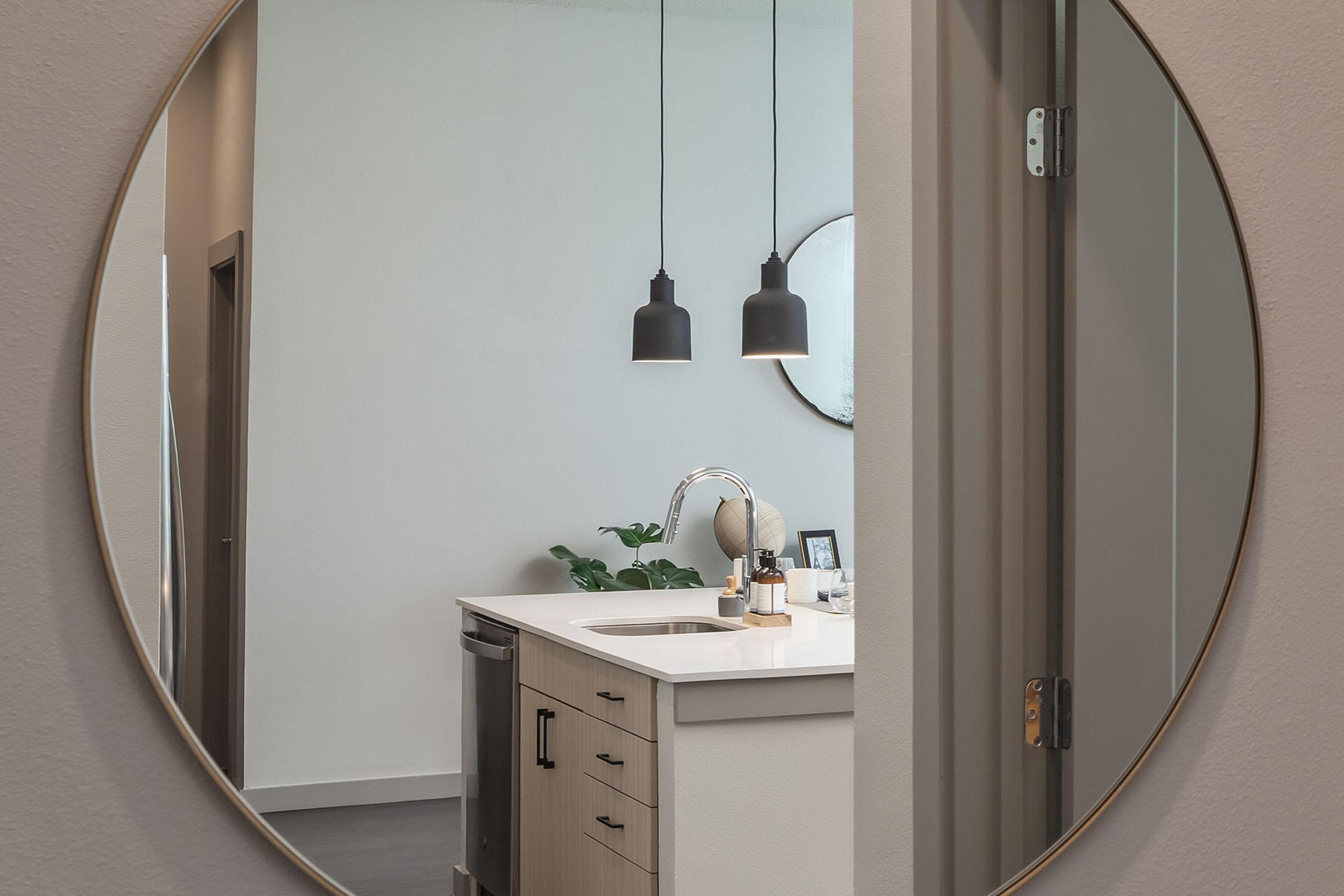 A round mirror reflecting a modern kitchen space with a white countertop, black pendant lights hanging above, and a small plant on the counter. The image features a door frame leading to the kitchen area, showcasing a clean, contemporary design.