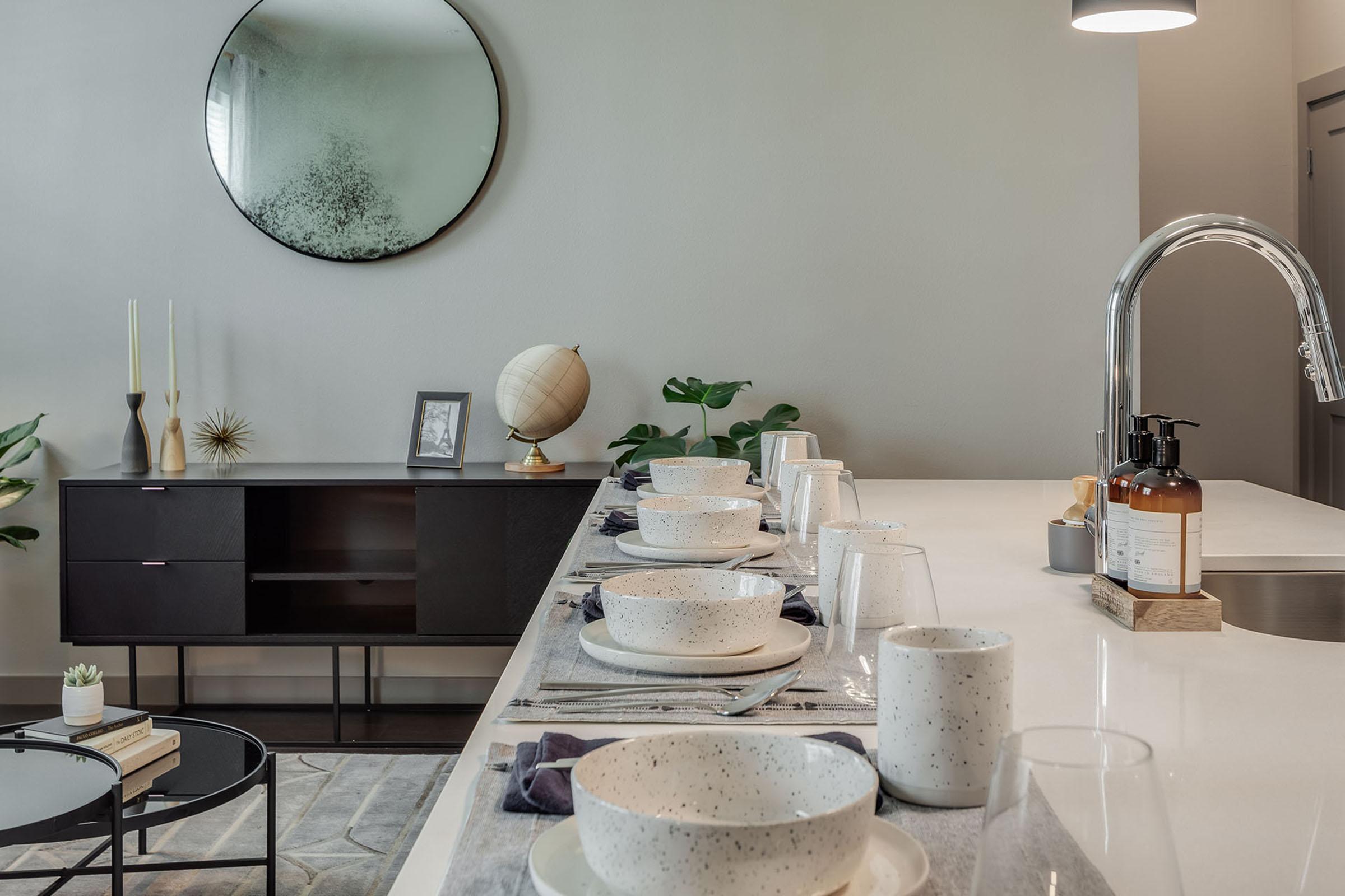 A modern dining area featuring a long table set with speckled white bowls, glasses, and navy napkins. In the background, a round mirror and a globe decorate the wall, alongside potted plants. A dark sideboard holds decorative objects, creating an inviting and stylish atmosphere.