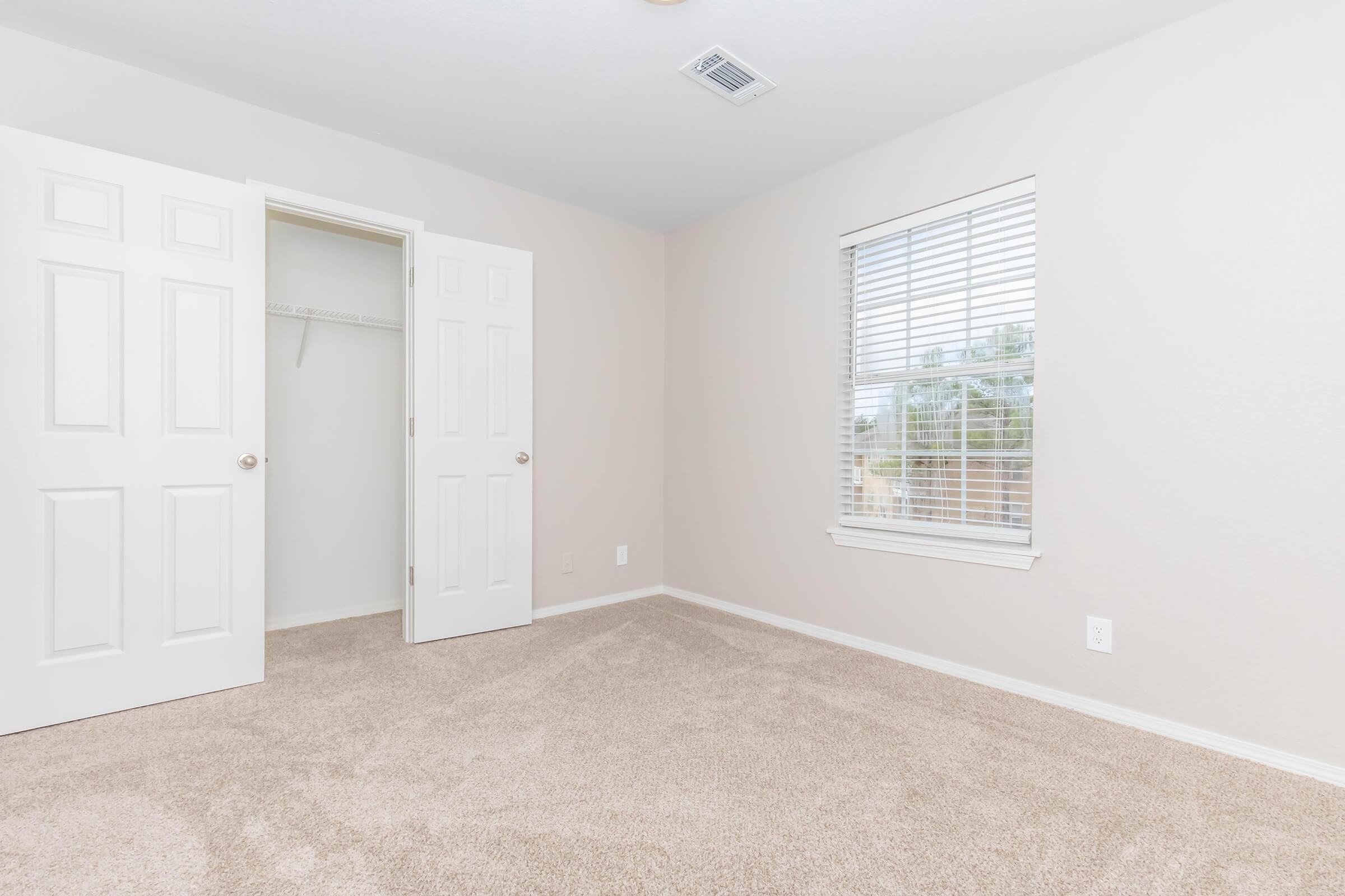 A clean, empty bedroom featuring light beige walls, a closed door leading to a closet, and a window with white blinds. The floor is covered in soft beige carpet, creating a cozy atmosphere. Natural light enters the room, highlighting the neutral color scheme.