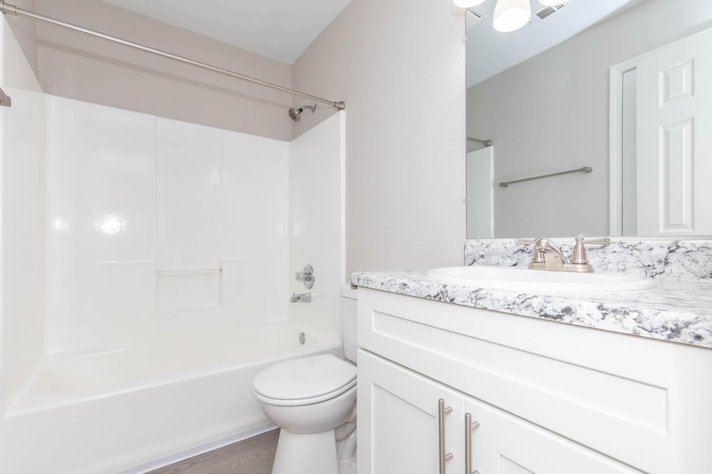 A clean and modern bathroom featuring a white bathtub, a countertop with a marble finish, a sink with silver faucets, and a light-colored wall. The room has a mirror, a light fixture above the sink, and a door in the background. The overall design is minimalistic and functional.