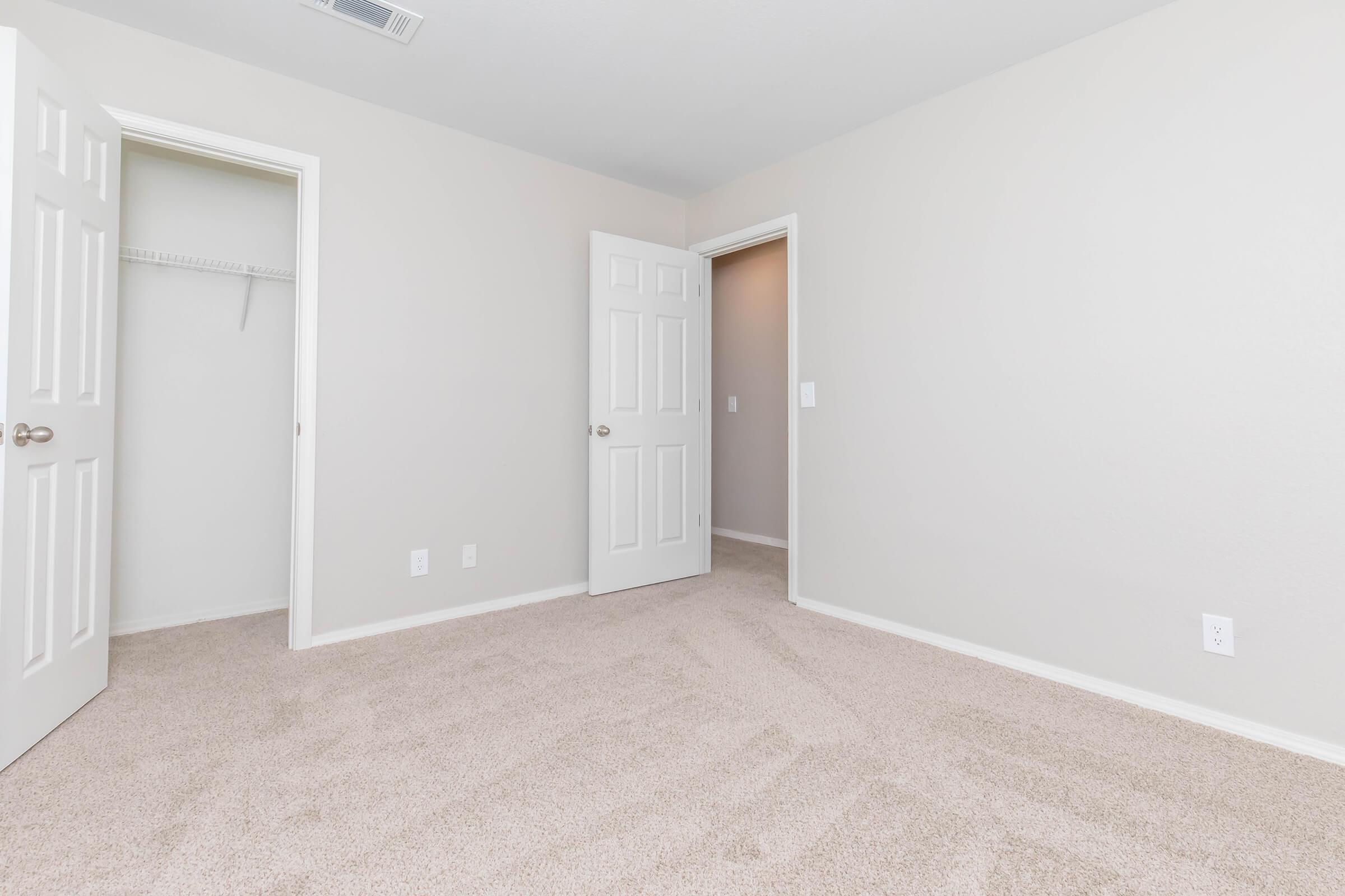 A spacious, empty room with light-colored walls and new carpet. Two doorways are visible, one leading to a closet and the other to a hallway. The room is well-lit and features neutral decor, making it suitable for various purposes.