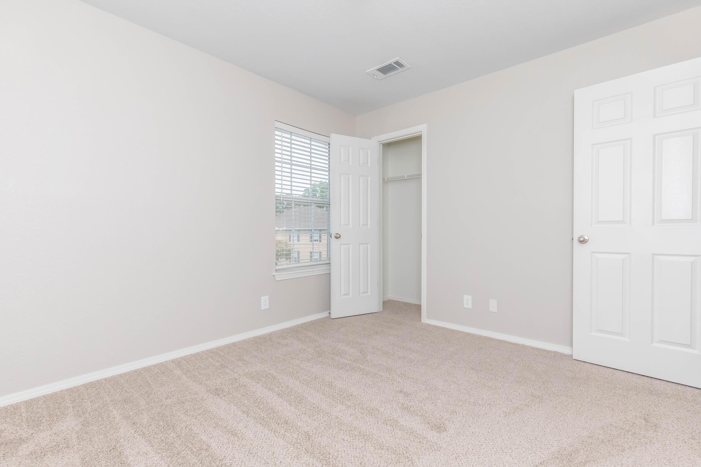 A sparsely furnished room with light beige walls and carpet. There is a window with blinds allowing natural light, and a closet with an open door. The opposite wall features a closed door. The space appears clean and minimalistic, ideal for various uses or decoration.