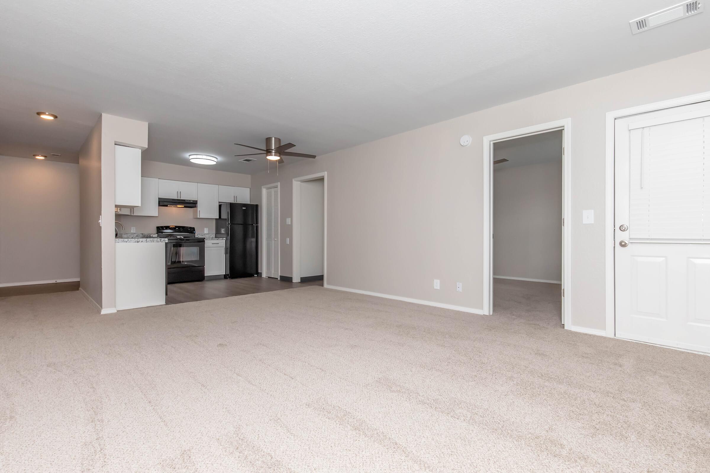Spacious living area with light-colored walls and carpet. A modern kitchen is visible in the background, featuring stainless steel appliances and a granite countertop. Doorways lead to additional rooms, and a front door is seen on the right. Ceiling fan and recessed lighting add to the ambiance.