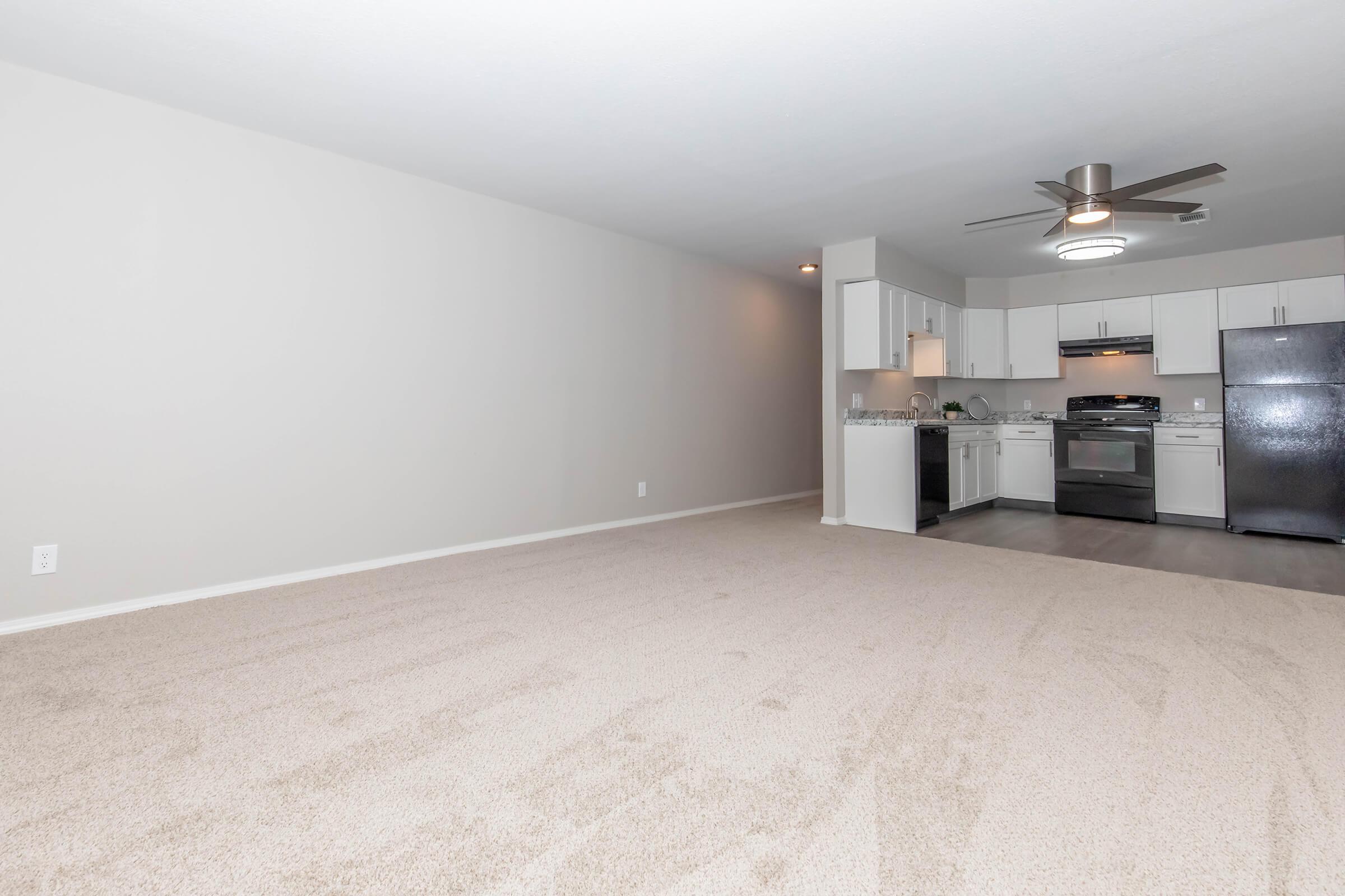 Spacious, well-lit room with light beige carpet, leading to a modern kitchen area. The kitchen features white cabinets, stainless steel appliances, and a ceiling fan. Wall is painted light gray, enhancing the open and airy feel of the space. Ideal for comfortable living or entertaining.