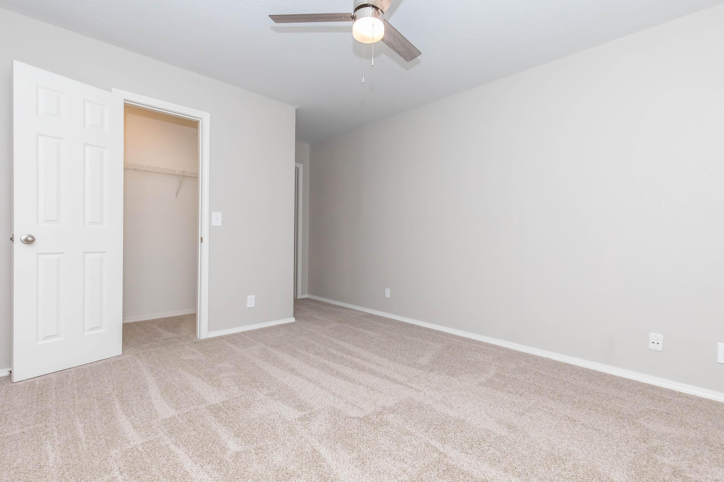 A well-lit, empty room with light gray walls and plush beige carpet. A ceiling fan is mounted above, and there is an open closet door on the left, revealing a small storage space. The room has a clean, minimalist appearance, suitable for various uses.