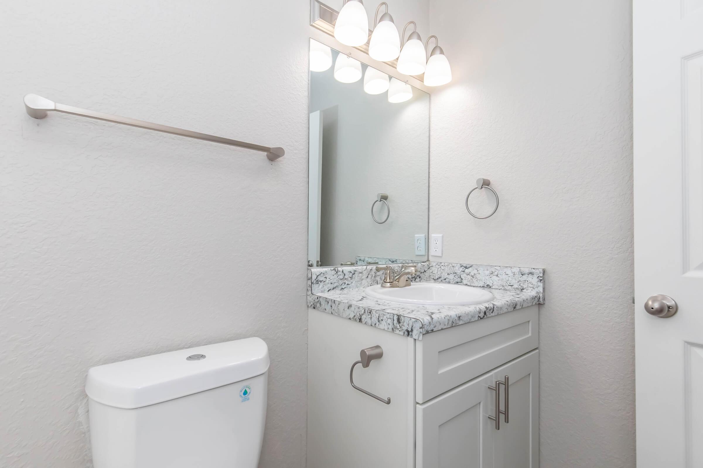 A modern bathroom featuring a white toilet, a gray and white granite countertop with a sink, a rectangular mirror above the sink, and five light fixtures. The walls are painted in a light color, and there is a towel bar and a towel ring on the wall.