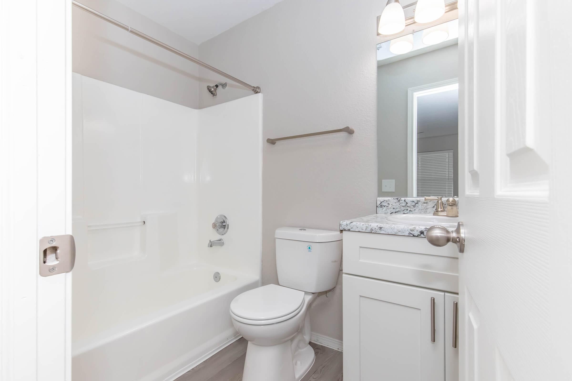 A clean and modern bathroom featuring a bathtub with a shower, a toilet, and a vanity with a marble countertop. The walls are painted in a light color, and there is a mirror with overhead lighting above the sink. The floor is tiled, contributing to the contemporary look of the space.