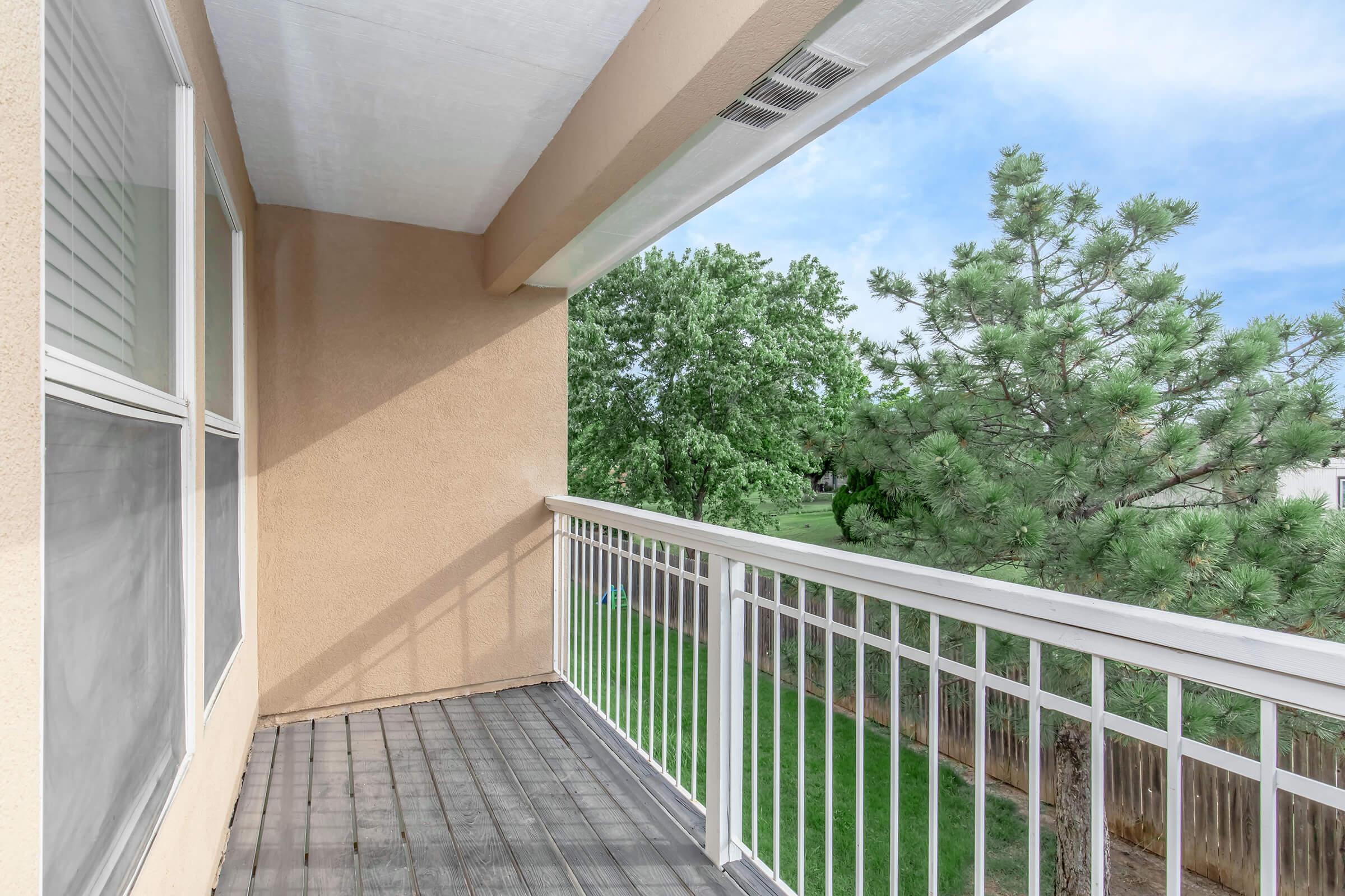 Balcony view featuring a wooden floor, white railing, and tan walls. In the background, there's a green tree and a grassy area, under a clear blue sky with scattered clouds. The setting suggests a serene outdoor space.