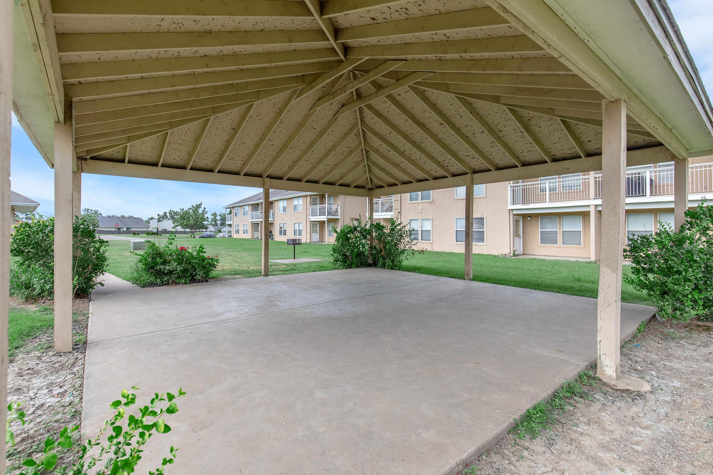 A spacious, covered outdoor pavilion with a peaked roof, featuring a concrete floor surrounded by green grass and small bushes. In the background, residential buildings are partially visible. The scene is bright and inviting, suitable for gatherings or relaxation.