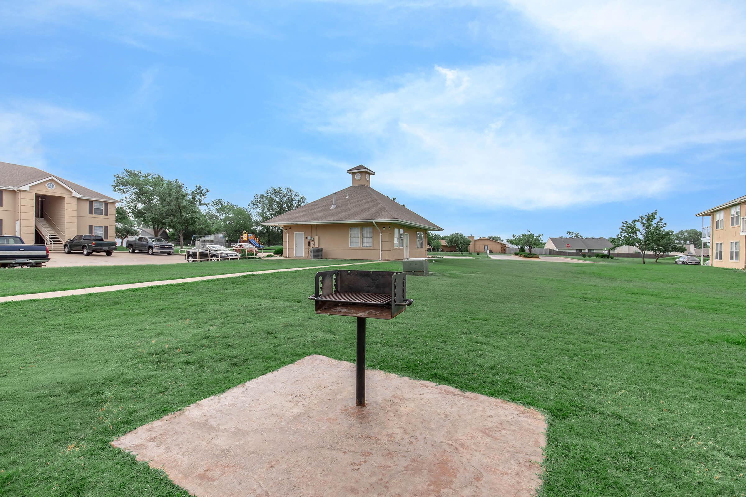 A grassy outdoor area featuring a single charcoal grill on a concrete slab, surrounded by well-maintained lawns. In the background, there are residential buildings and trees, with a clear blue sky above.