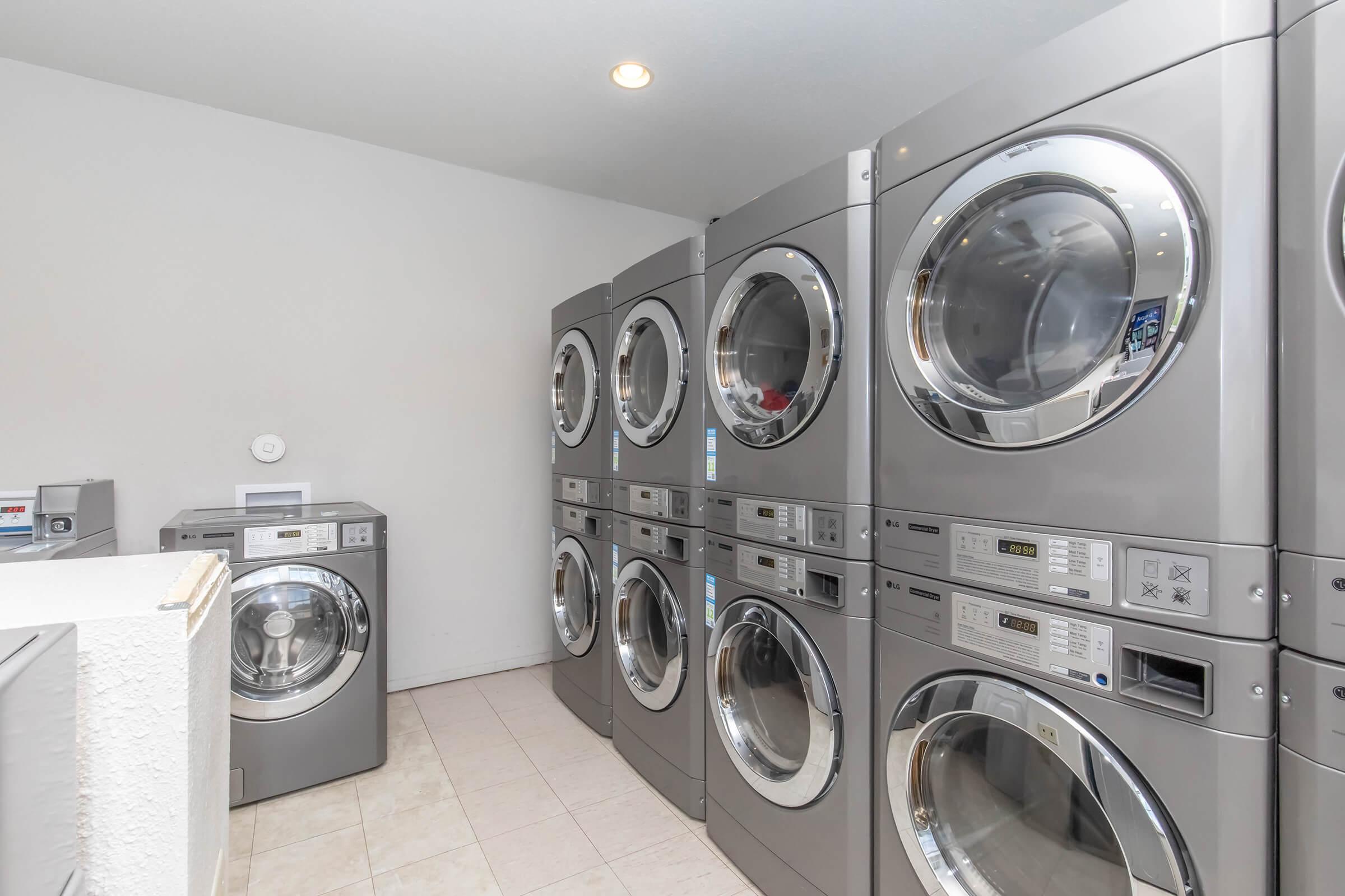 A laundry room featuring several stacked washing machines and dryers in a row, with a light-colored tiled floor and a wall painted in neutral tones, creating a clean and modern atmosphere. There is also a small area for folding laundry on the left.