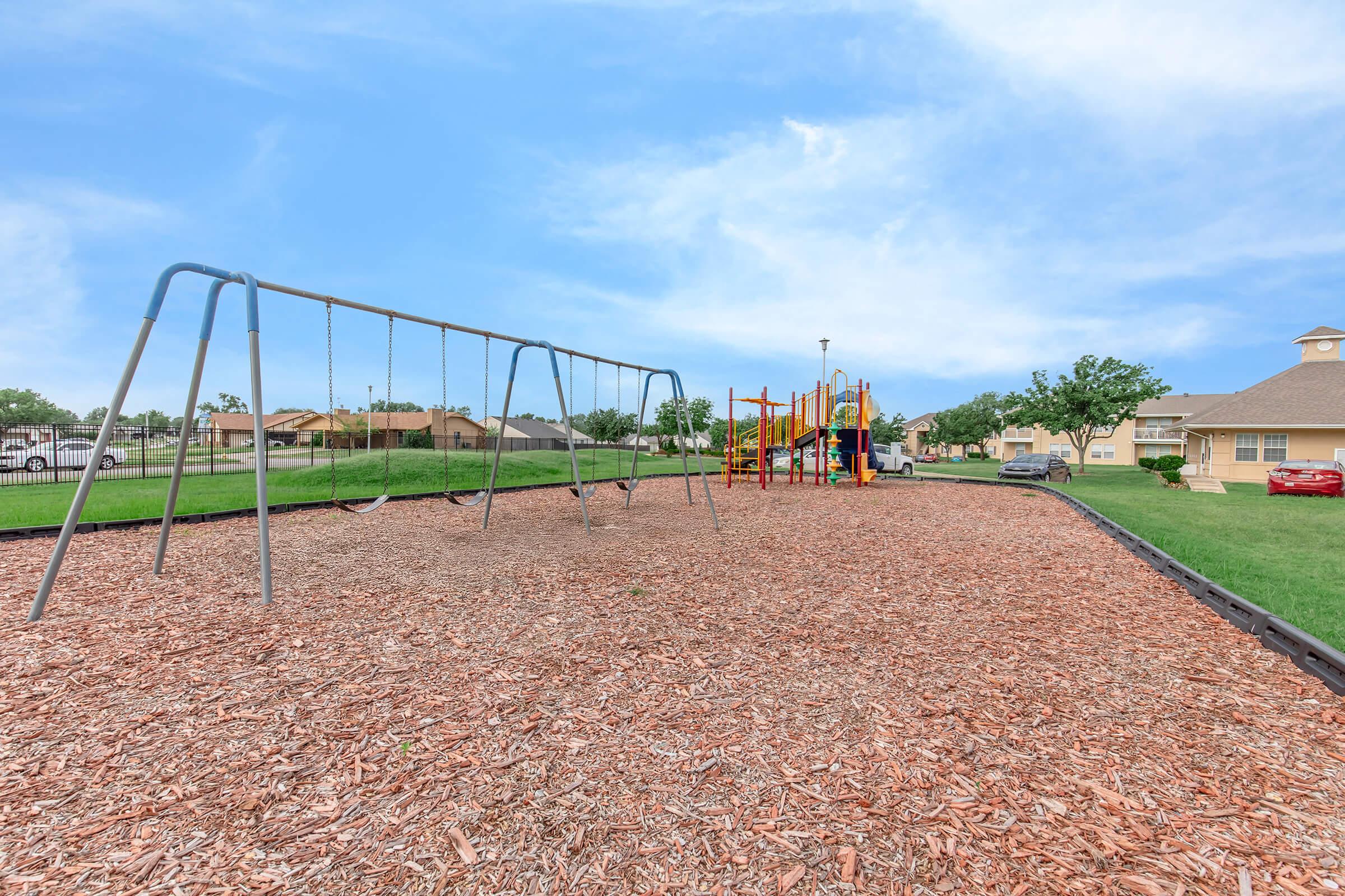 A playground scene featuring a set of swings on a bed of wood chips, with a colorful play structure in the background. Green grass surrounds the playground, and residential buildings can be seen in the distance under a clear blue sky.