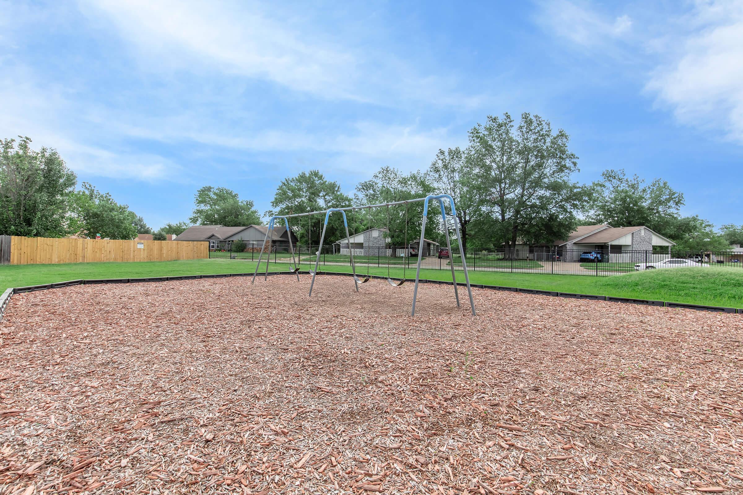 A playground scene featuring a swing set on a bed of wood chips. In the background, there are residential homes and lush green trees under a partially cloudy sky. The area is open and inviting, suggesting a community space for outdoor activities.