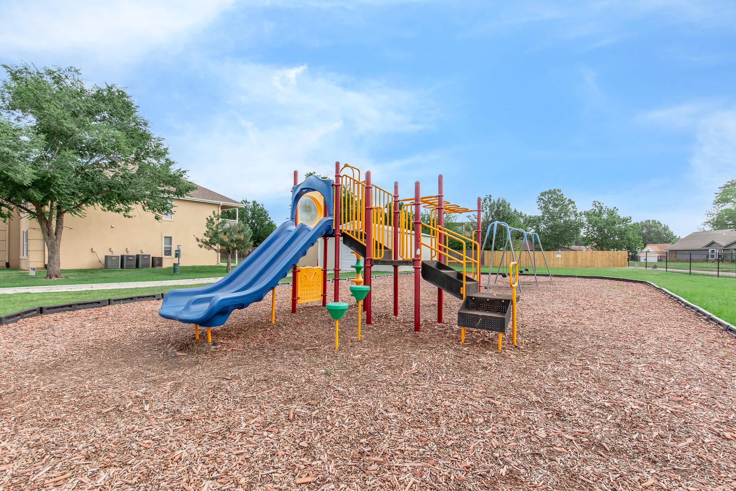 Colorful playground equipment with blue and yellow slides, monkey bars, and swings, set in a grassy area surrounded by mulch. In the background, residential buildings and trees are visible under a clear blue sky.