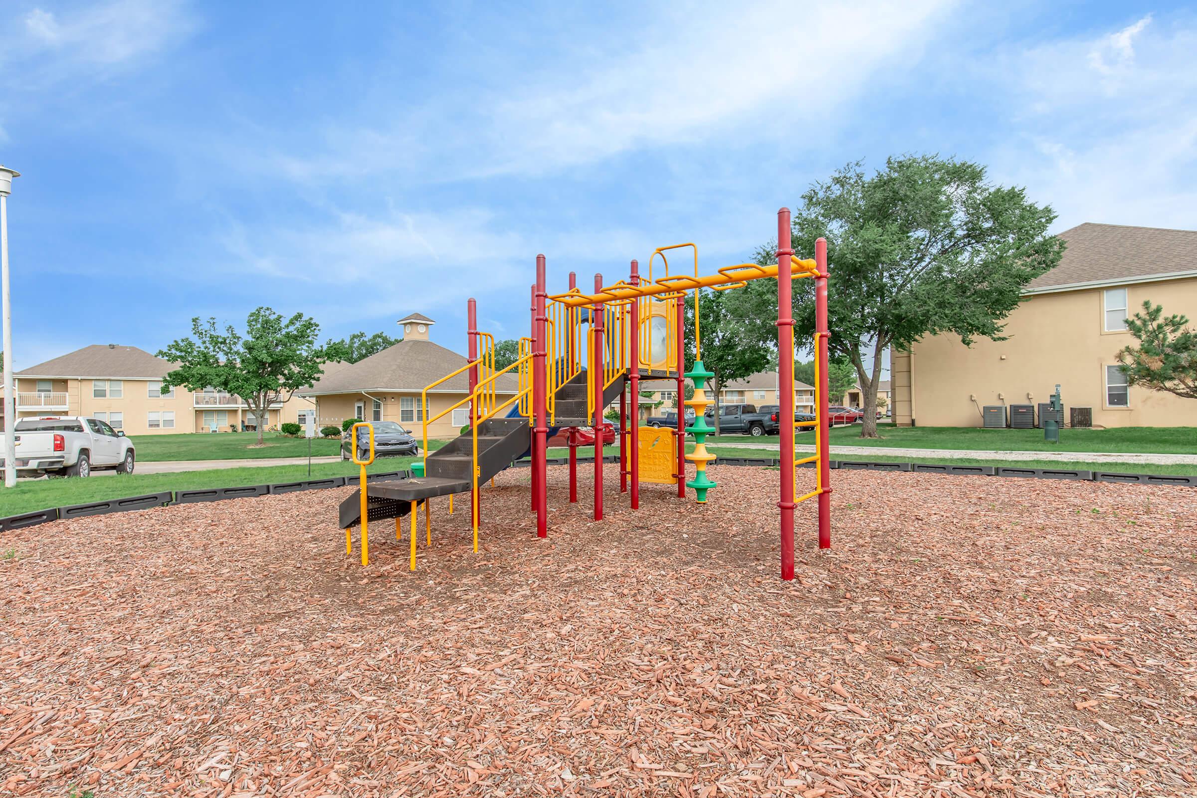 A colorful playground structure with yellow and red equipment, including slides and climbing elements, situated on a bed of wood chips. Green trees and residential buildings are visible in the background under a blue sky.