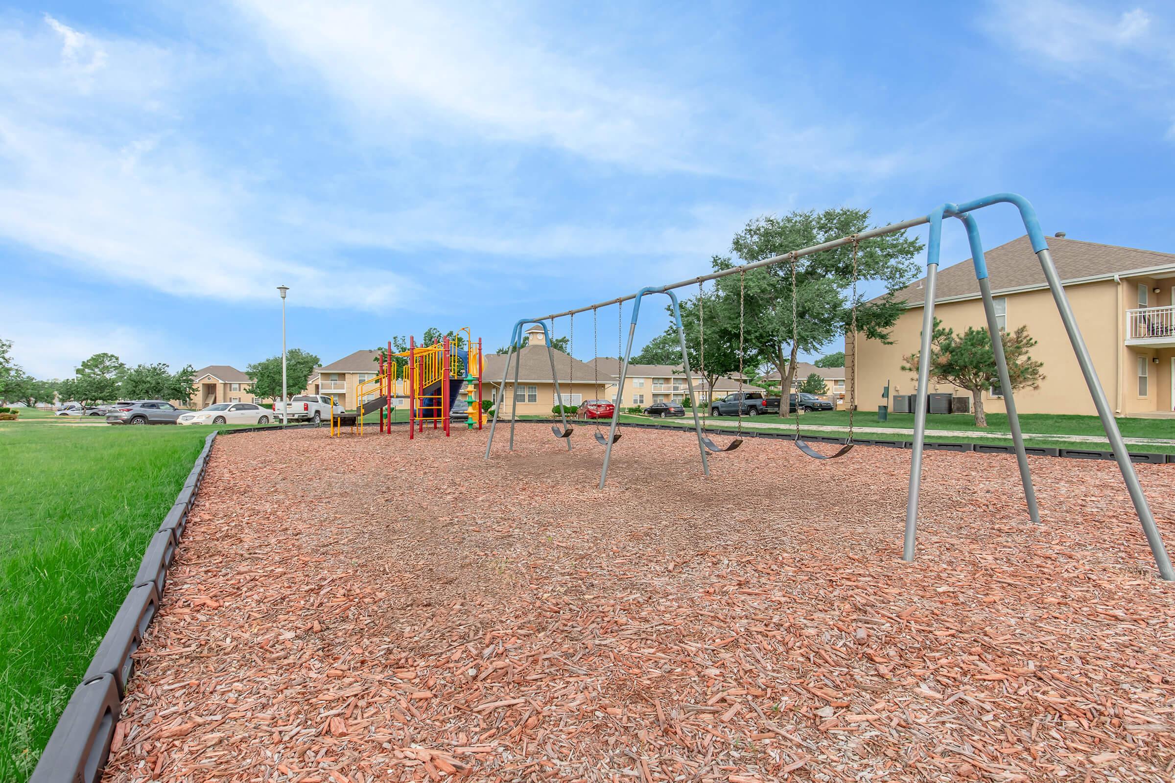 A playground featuring swings, a play structure, and soft mulch ground cover, set in a grassy area near residential buildings. The sky is blue with scattered clouds, and cars are parked nearby.