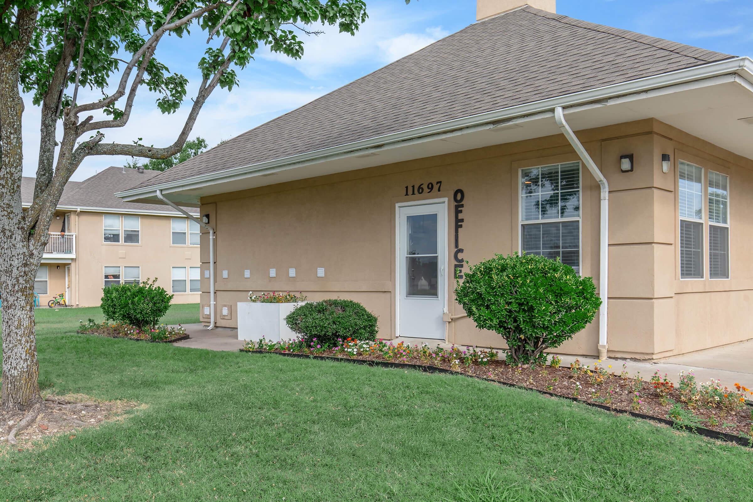 A beige office building with a sloped roof, located in a landscaped area with green grass and flowers. The building features a white door and several windows, with the number "11697" displayed on the wall. A tree is visible nearby, providing shade. The setting is bright and well-maintained.