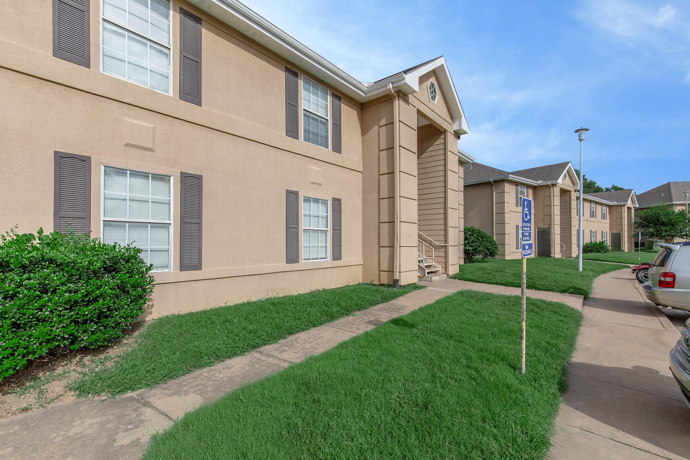 A view of a beige multi-unit apartment building with white windows and dark shutters. The entrance features a small staircase, surrounded by neatly trimmed green grass. Parking spaces are visible in front, and the sky is clear with a few clouds.