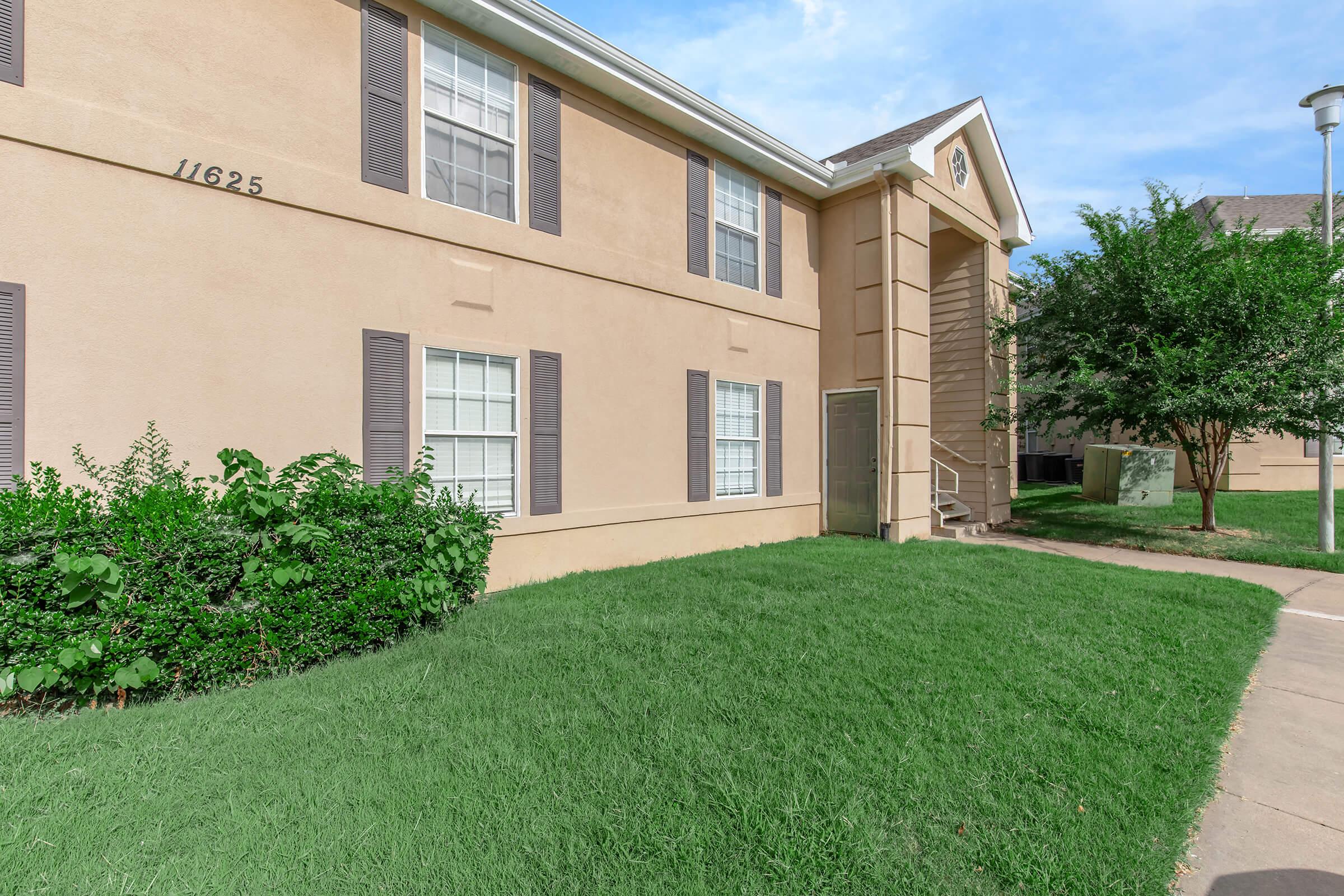 A two-story beige apartment building with white windows and dark shutters. In the foreground, there is a green lawn with bushes and a walkway leading up to the entrance. The sky is partly cloudy, creating a bright atmosphere.