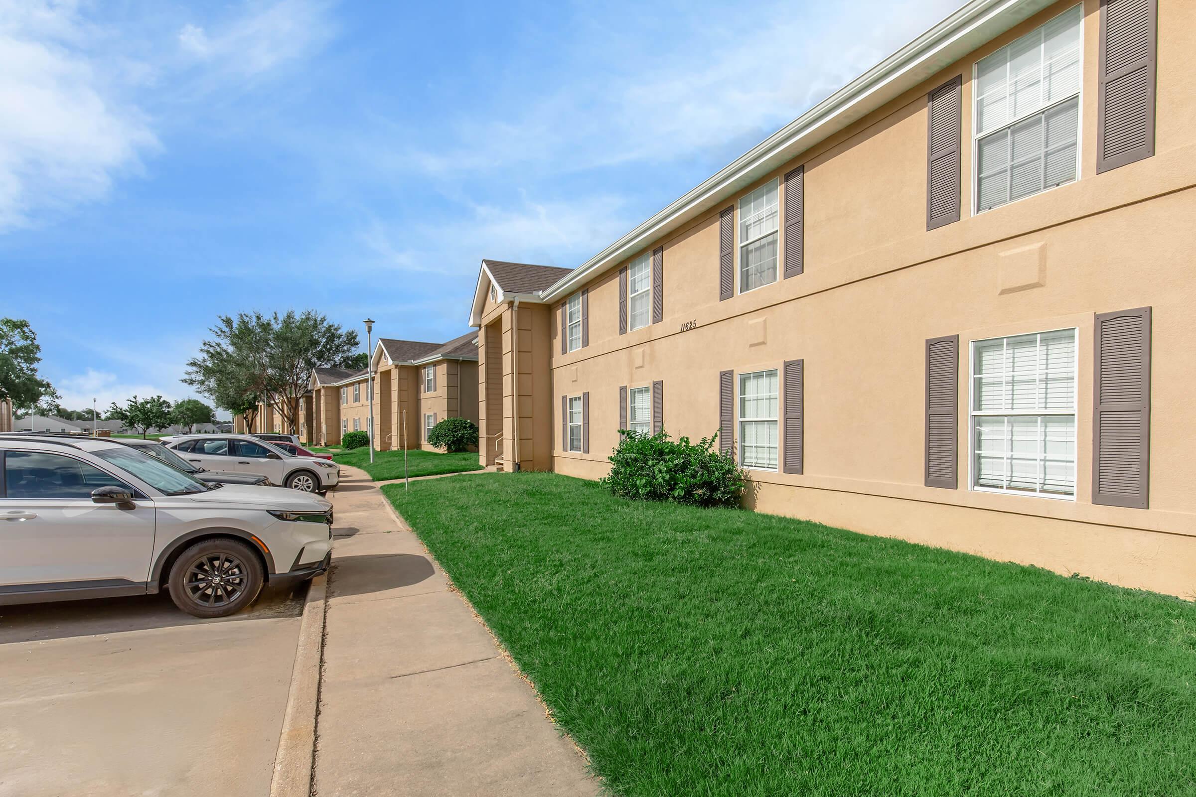 A view of a residential apartment building with tan-colored exterior walls and several windows. In the foreground, there is a parking lot with cars parked along the side, and a well-maintained grass area. The sky is clear with a few clouds.