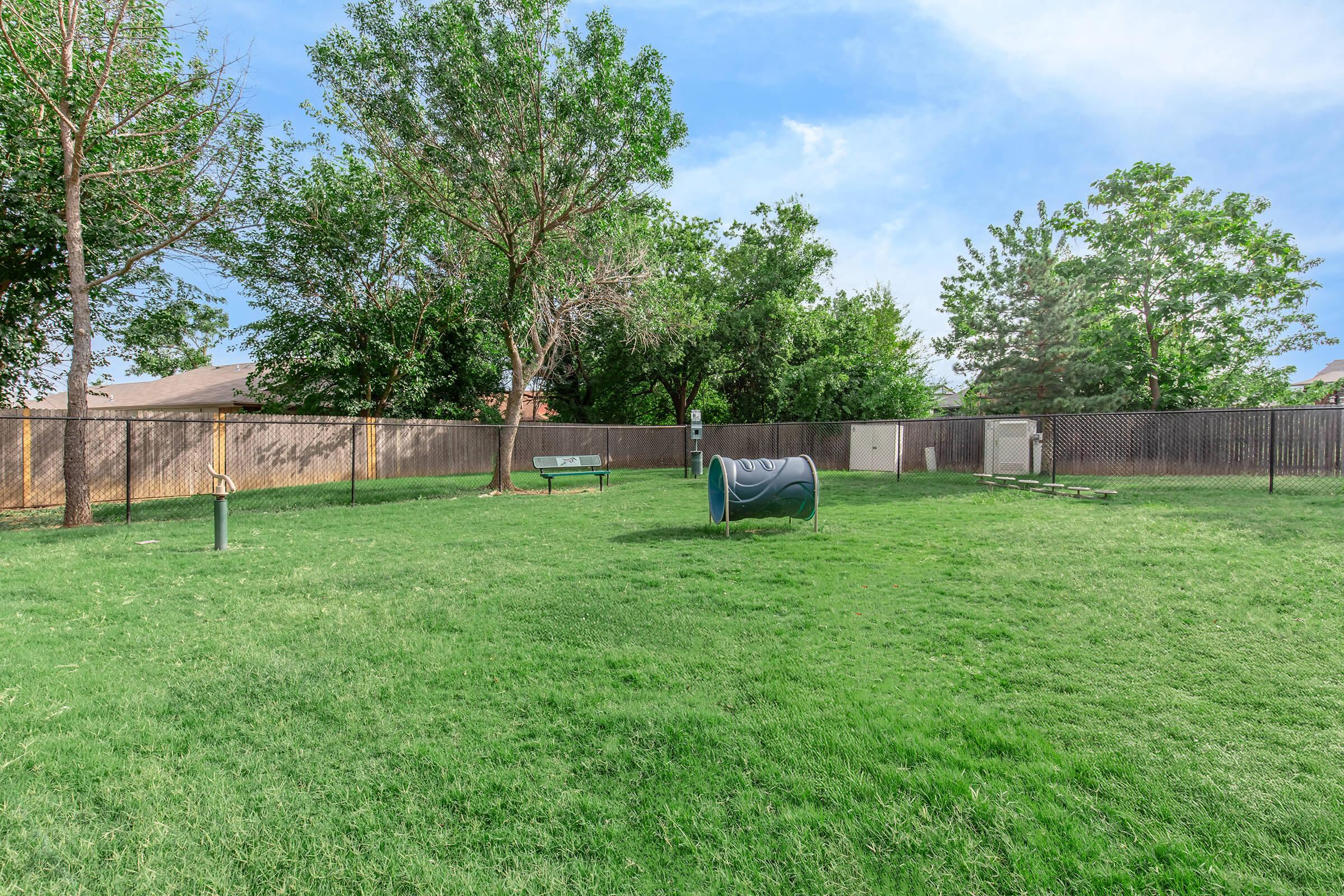 A spacious, fenced dog park featuring green grass, trees, and benches. Central to the area is a dog waste dispenser, with a few agility equipment pieces nearby. The park is well-maintained, with clear blue skies in the background, creating a welcoming environment for pets and their owners.