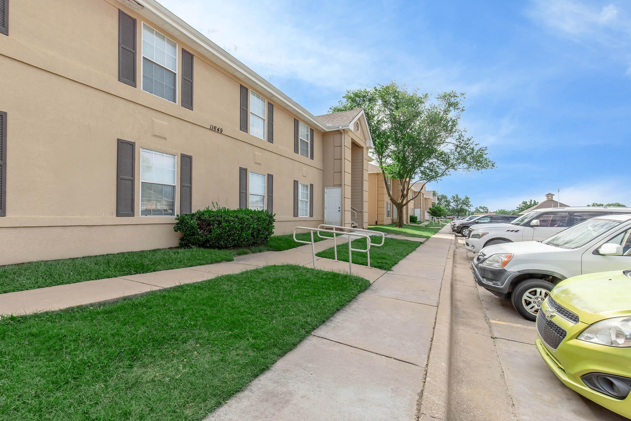 A well-maintained apartment complex with beige exterior walls, featuring several windows and shrubs. A concrete sidewalk leads to the entrance, with a railing for accessibility. Several parked cars line the parking lot on the right, and a clear blue sky is visible in the background.