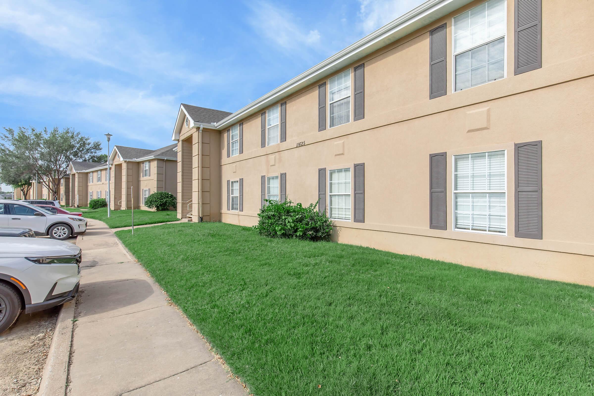 Exterior view of a multi-unit residential building with tan siding and white windows. The building features a well-maintained lawn and parking area with cars. Blue sky and scattered clouds are visible in the background. Pathway leading up to the entrance.
