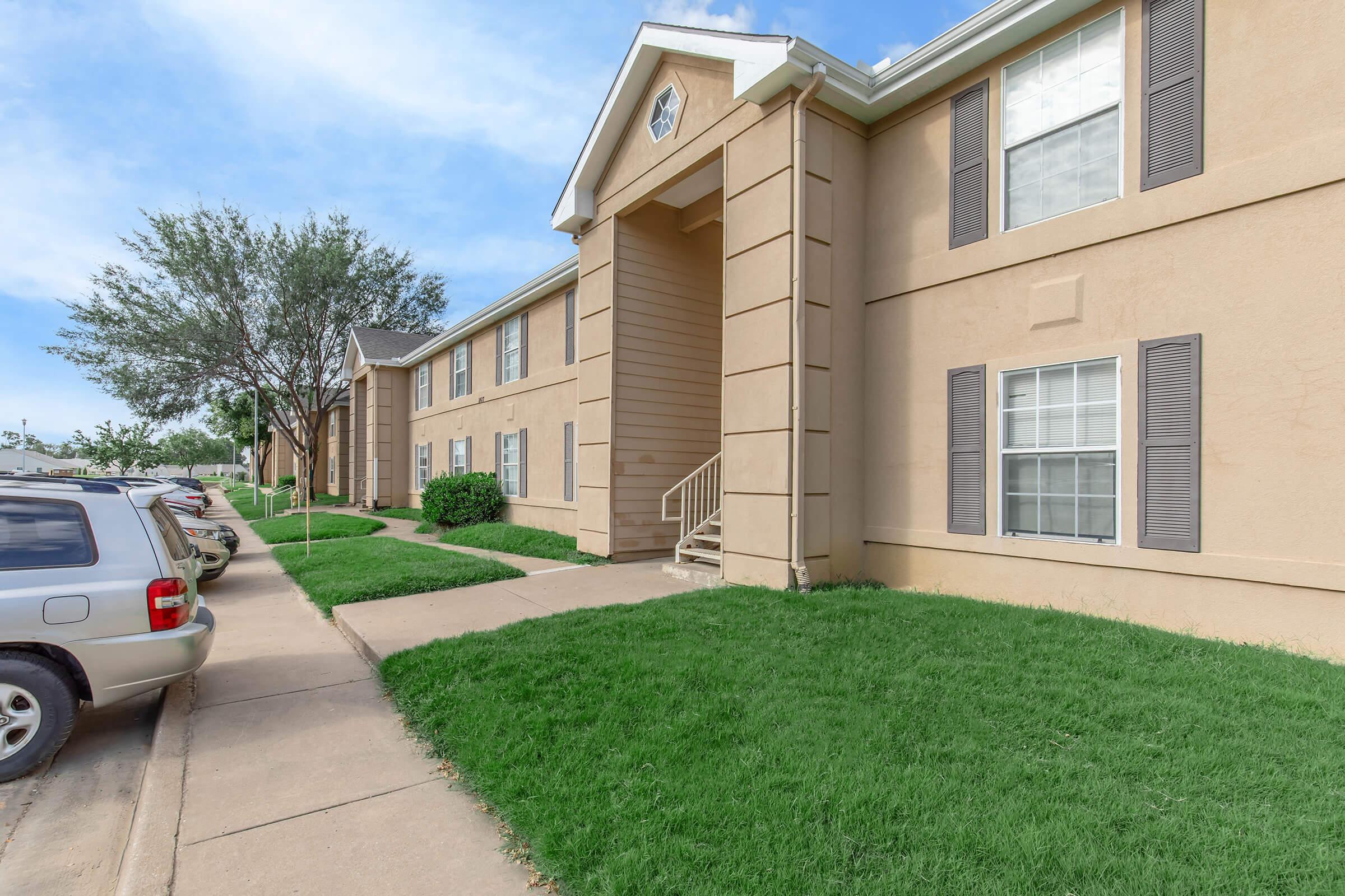 An exterior view of a two-story apartment building featuring a light beige facade, green grass, and a sidewalk. Several parked cars are visible nearby, along with trees in the background and a clear blue sky above.