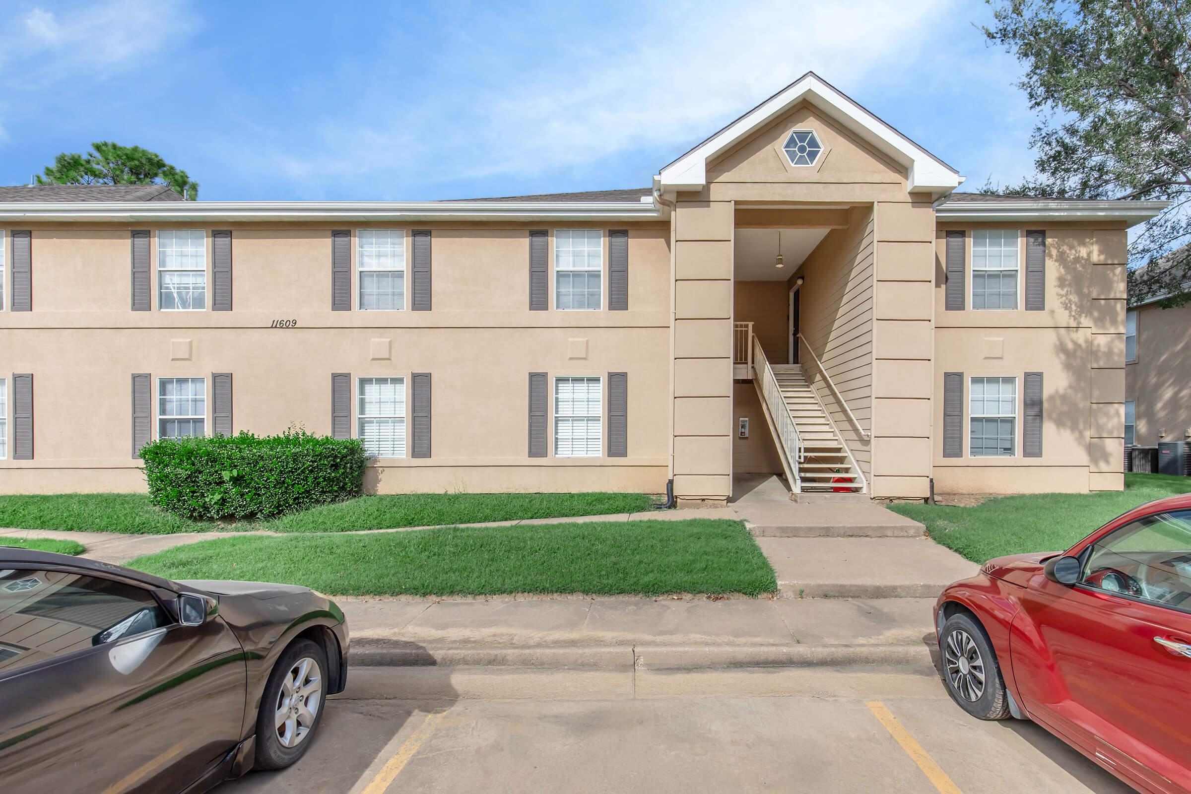 A tan two-story apartment building with multiple windows and brown shutters, surrounded by green grass. A staircase leads to the front entrance, and there are two parked cars—one black and one red—in the foreground, with a clear blue sky above.
