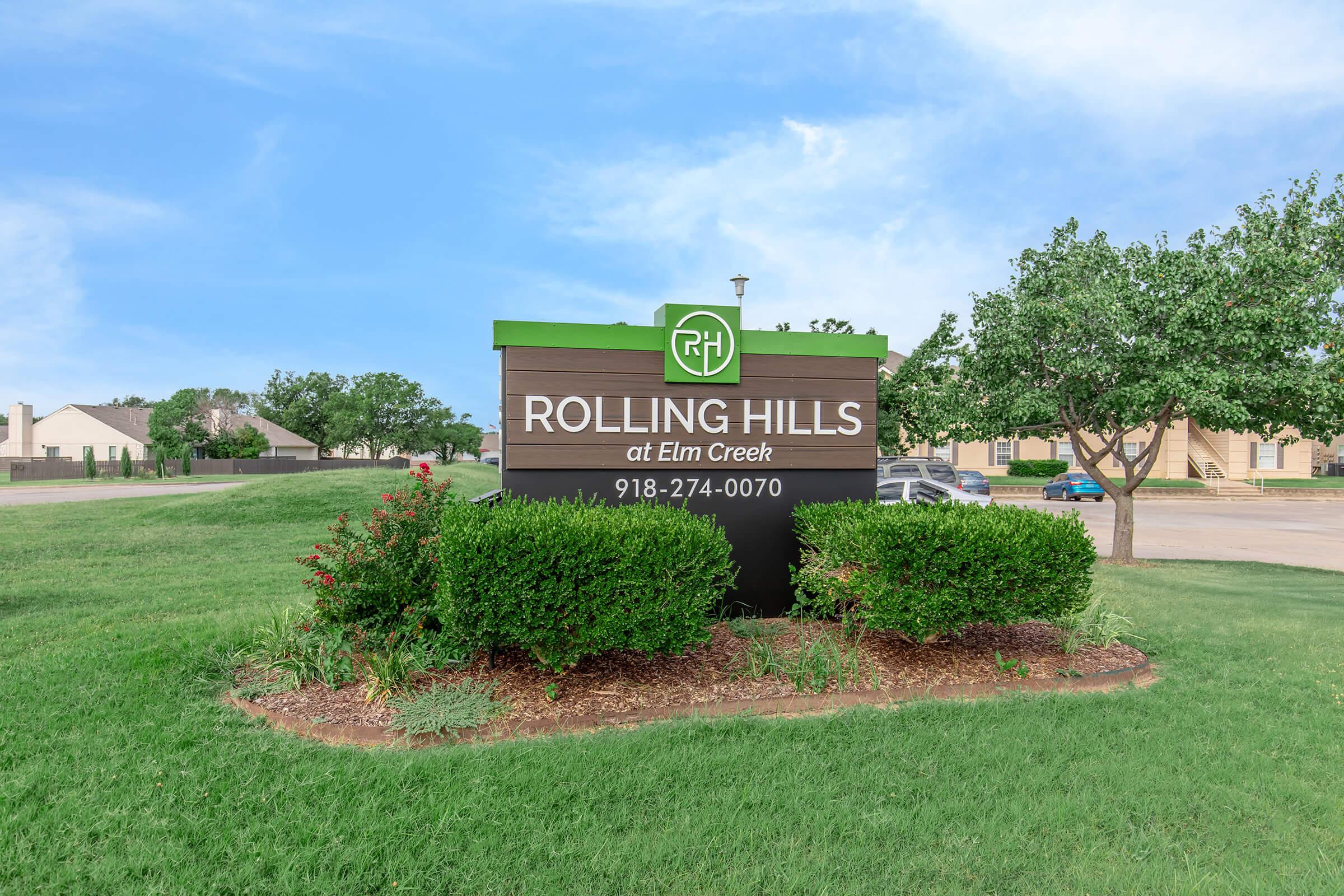 A sign for "Rolling Hills at Elm Creek," featuring the name prominently displayed along with contact information. The sign is surrounded by well-manicured shrubs and grass, with a clear blue sky in the background.