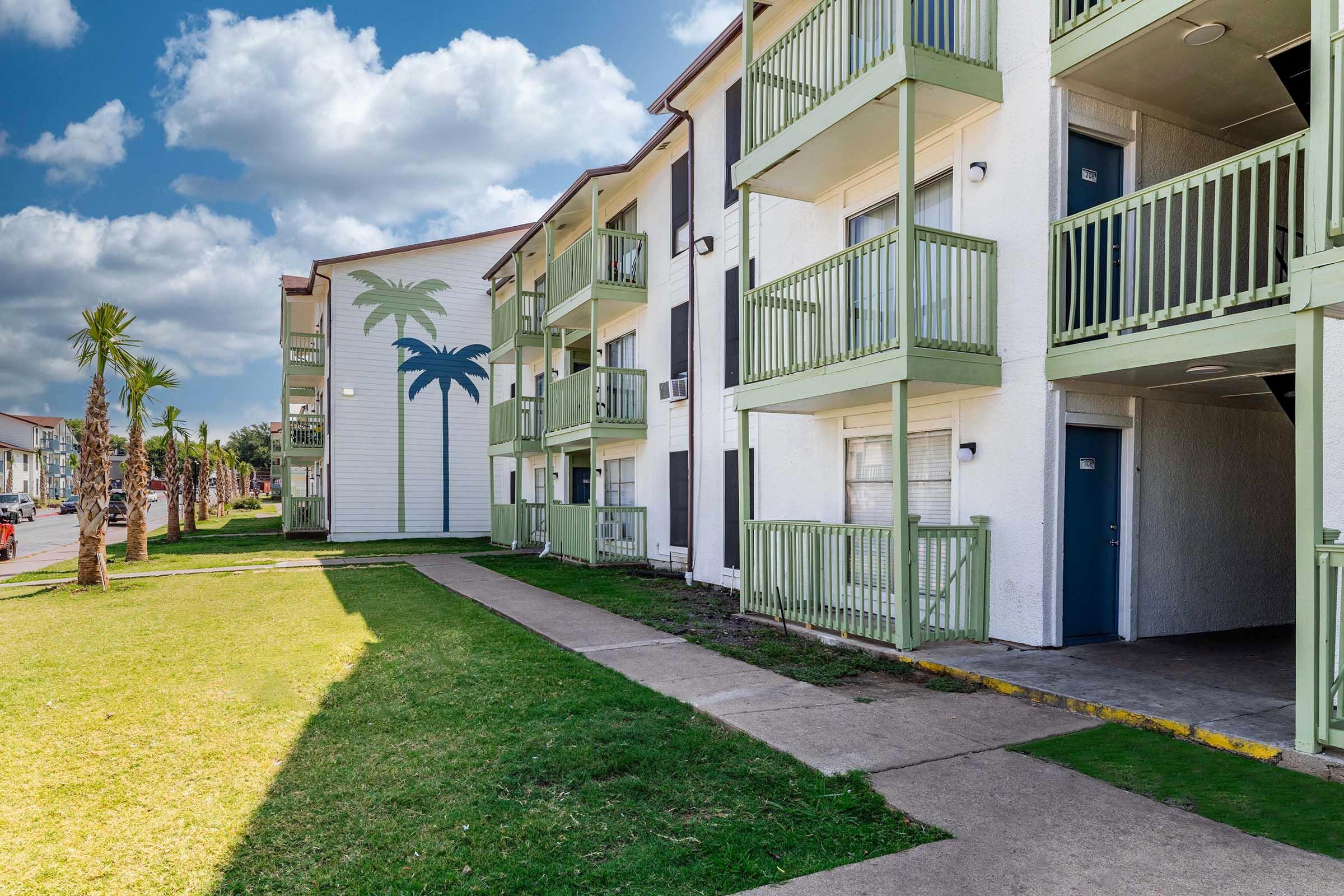 A view of a multi-story apartment complex featuring light-colored buildings with green balconies. The foreground includes a paved walkway and grassy area, while a mural of palm trees adds a tropical touch. The background shows a cloudy sky, enhancing the overall aesthetic of the setting.