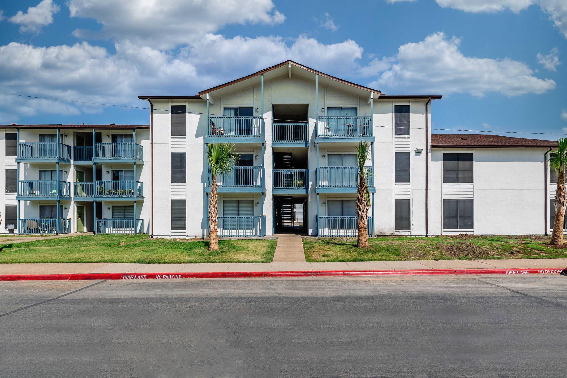 A three-story apartment building with a white exterior and blue balconies, surrounded by green grass and palm trees. The building features a central staircase, and the photo is taken on a sunny day with blue skies and some clouds. A paved road runs in front of the building.