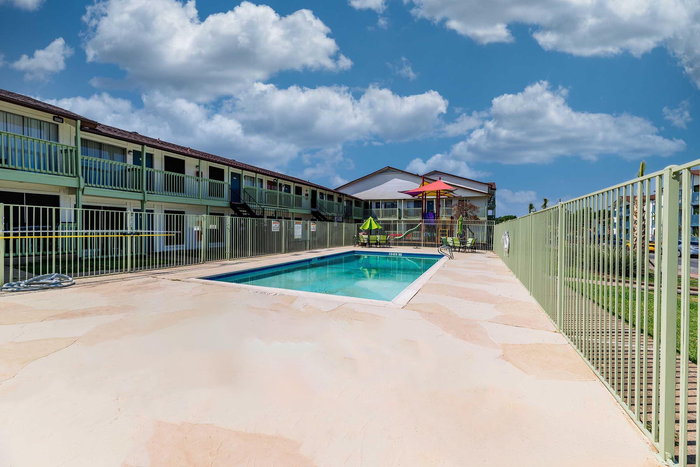 A clear swimming pool surrounded by a fence, with lounge chairs and an umbrella nearby. In the background, there is a low-rise building with multiple rooms, under a bright blue sky with fluffy white clouds. The area appears welcoming and well-maintained, perfect for relaxation.